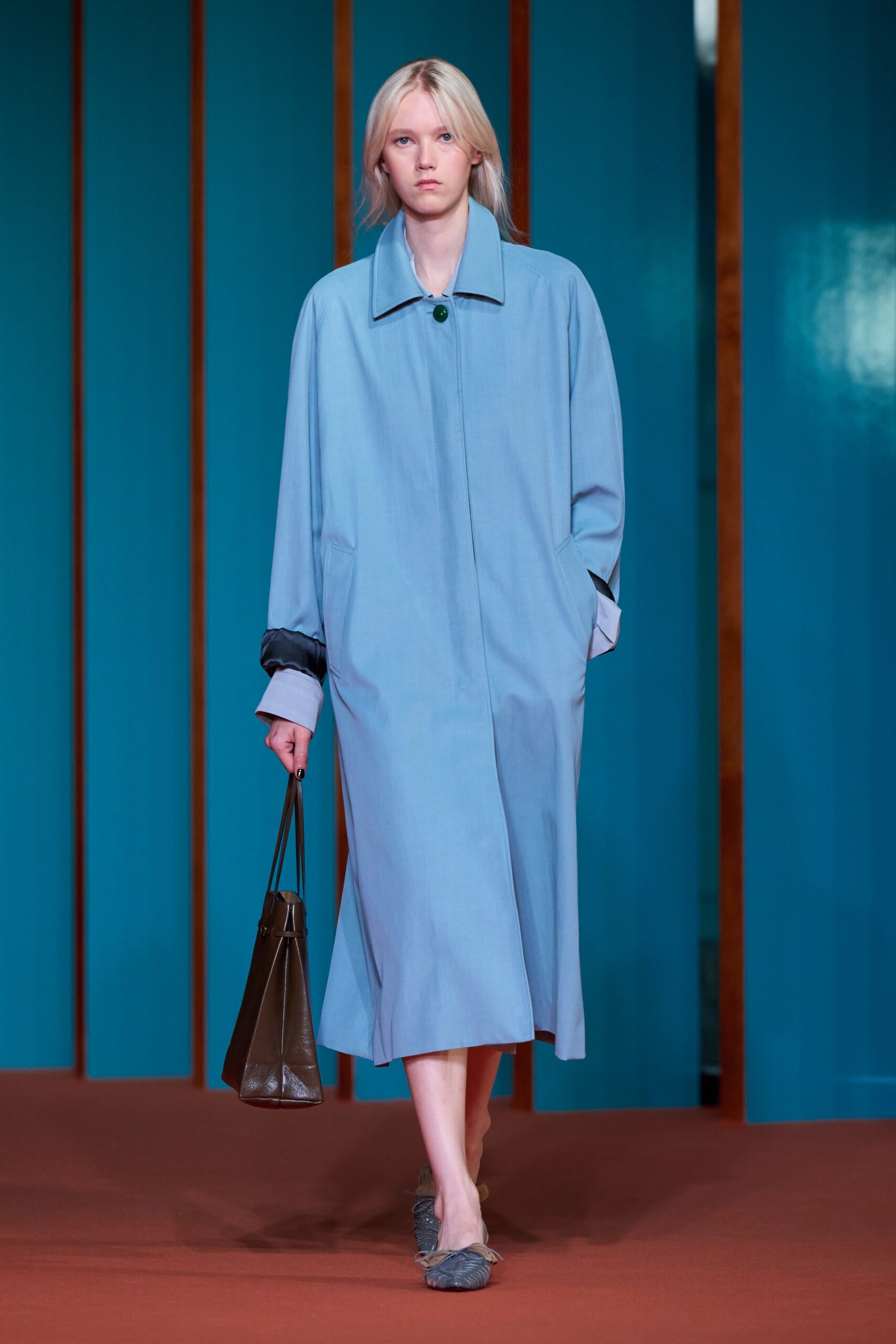Powder-blue coatdress in fluid cloth, cut with dropped shoulders and one dark green button. White shirt cuffs turned back beneath charcoal satin sleeve linings. Deep brown leather bucket bag with long thin straps. Slate gathered flats tied with a pale cord bow. For crossing a quiet hotel lobby before breakfast is being cleared.