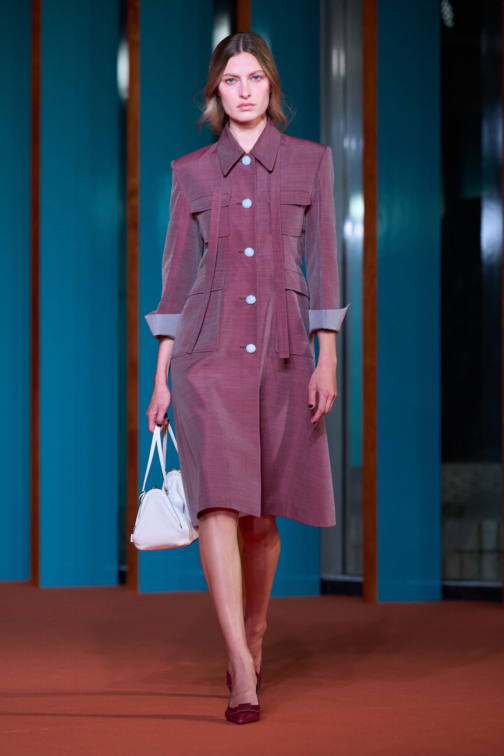 Dusty mauve coatdress in slubbed suiting, cinched with hanging self-tabs and pale round buttons. White leather top-handle bag with twin zipped lobes. Oxblood pumps with a squared bow across the vamp. For arriving early to a civic luncheon in a building with too much teal glass.