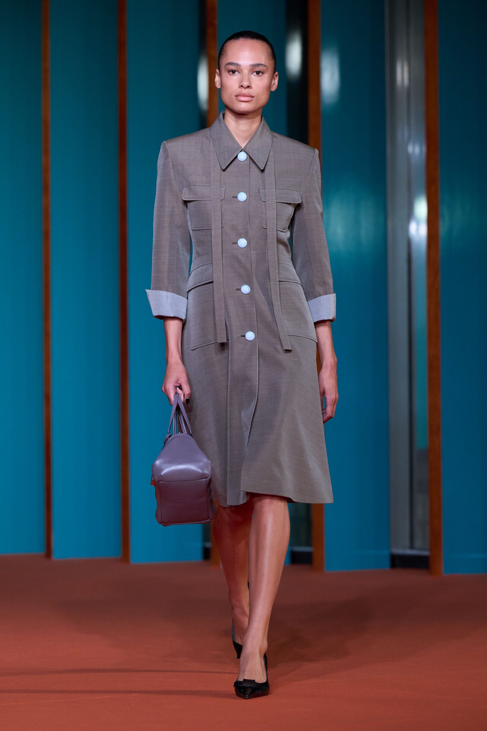 Taupe worsted coatdress with pale round buttons and long hanging plackets. Dusty mauve leather carryall, squared off at the base. Black pointed pumps with a small bow at the vamp. For signing papers in a municipal building after the lunch crowd clears.