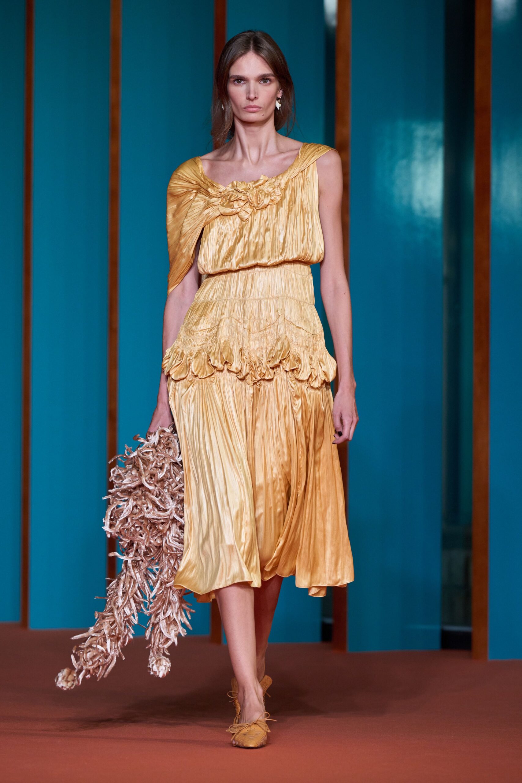 Honey satin dress in crushed pleats, rosettes knotted across one shoulder. Long tasselled carryall trailing pale curled strips like dried palm fiber. Ochre lace-up flats with a pinched square toe. For crossing a tiled courtyard after lunch, heat still sitting in the walls.