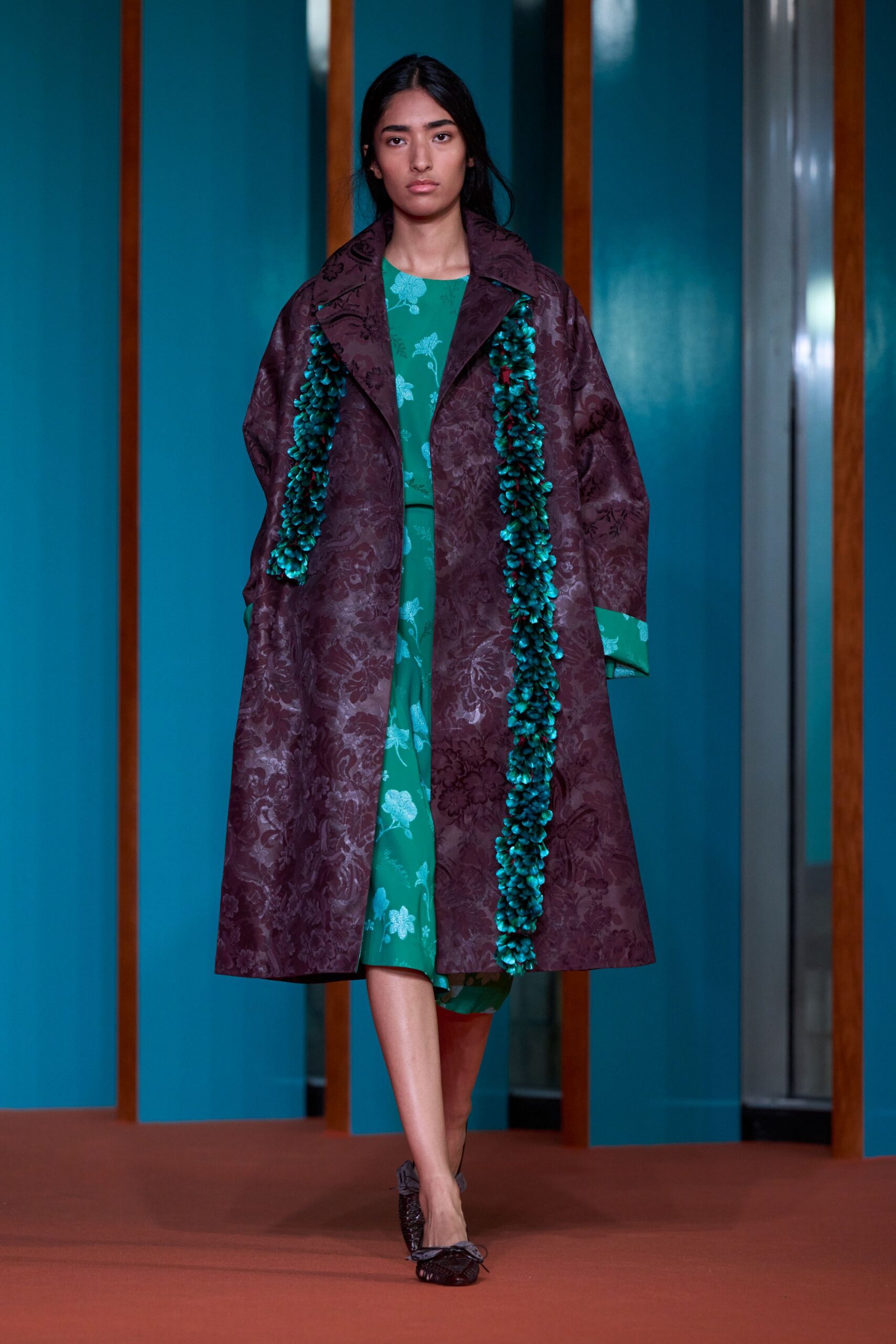Plum floral jacquard coat with thick teal petal garlands down the front. Emerald dress scattered with pale woven blossoms, hem peeking below the coat. Dark crinkled flats tied with a small bow. Crossing a tiled courtyard after rain, carrying petals back from a ceremony.