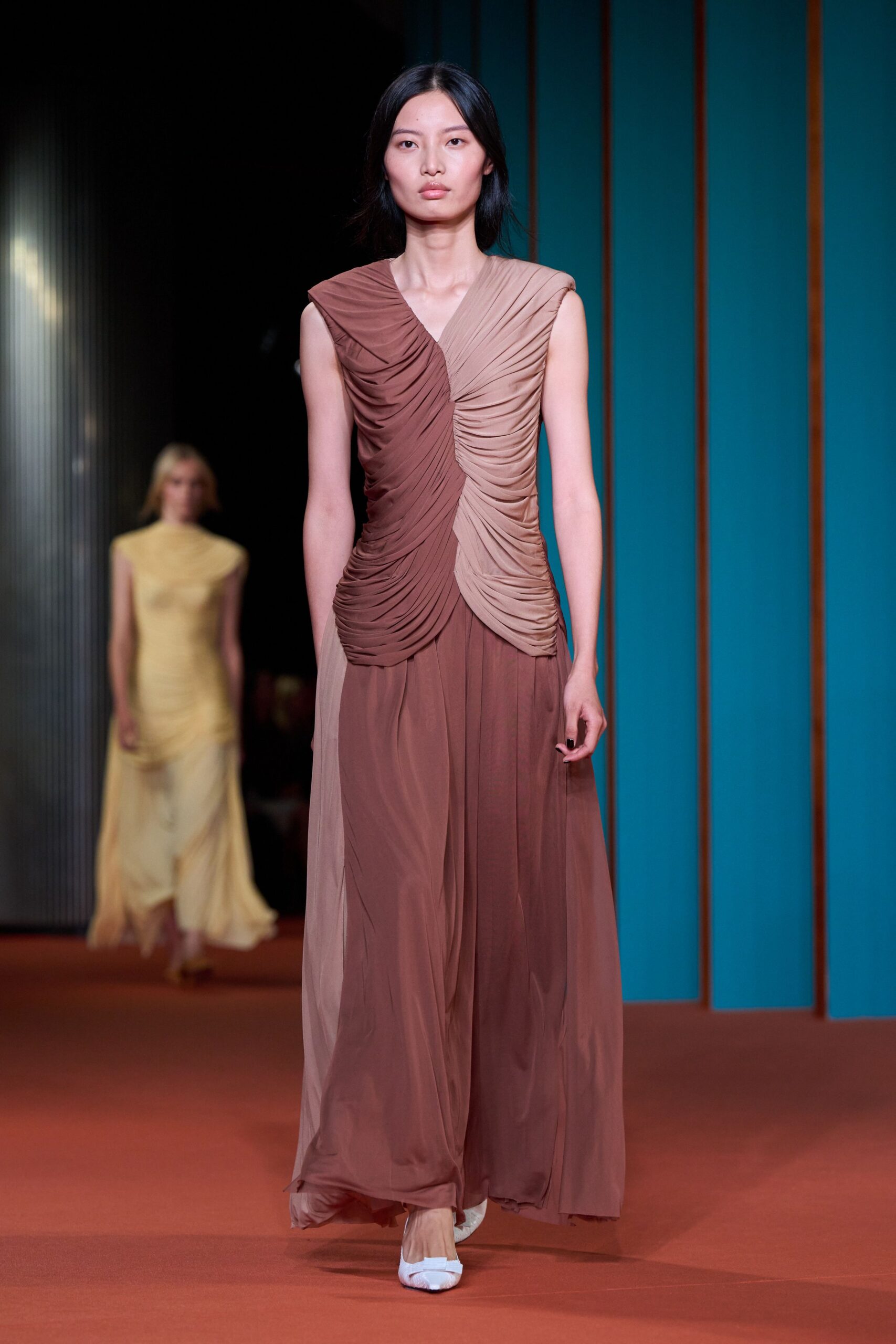 Sleeveless two-tone chiffon gown, densely ruched through the bodice and hips. White pointed flats peeking from the pooling hem. Crossing a tiled hotel atrium before the florist has finished unloading.