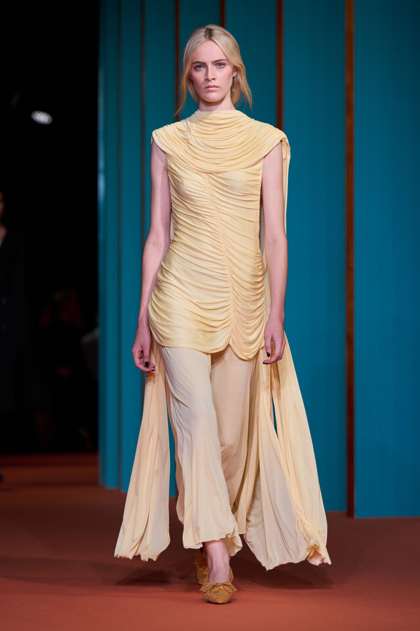 Buttercream jersey tunic densely ruched to a high cowl, one shoulder trailing. Pale georgette trousers cut wide and liquid under the asymmetric hem. Tan leather flats with gathered toes and loose wrap laces. Crossing a hotel courtyard at dawn before the fountains switch on.