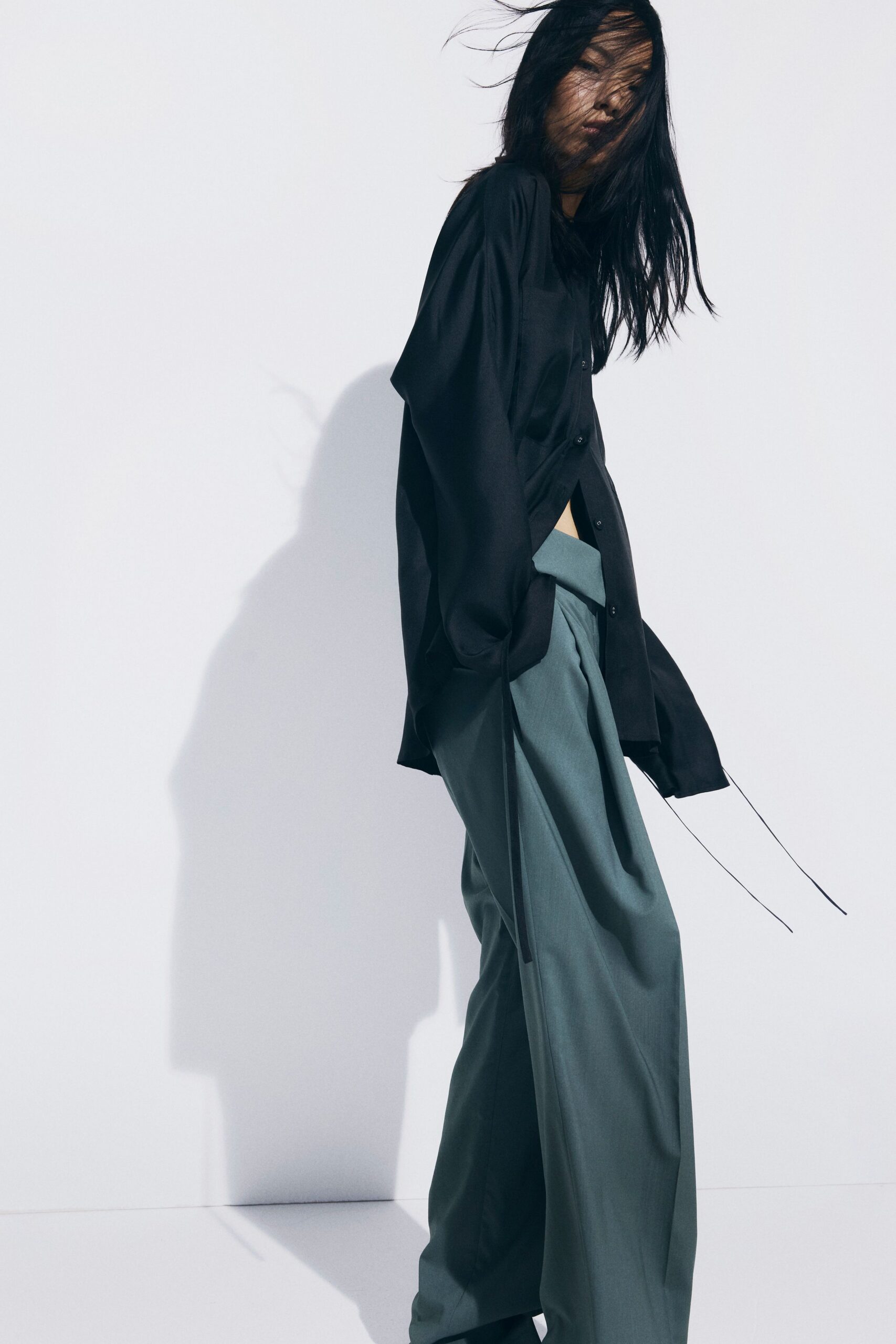 Black fluid shirt hanging open, with extra-long cords trailing from the cuff. Sage wide-leg trousers with a folded waistband and deep front pleats. Wind tunnel in a photo studio, somebody just opened the loading door.