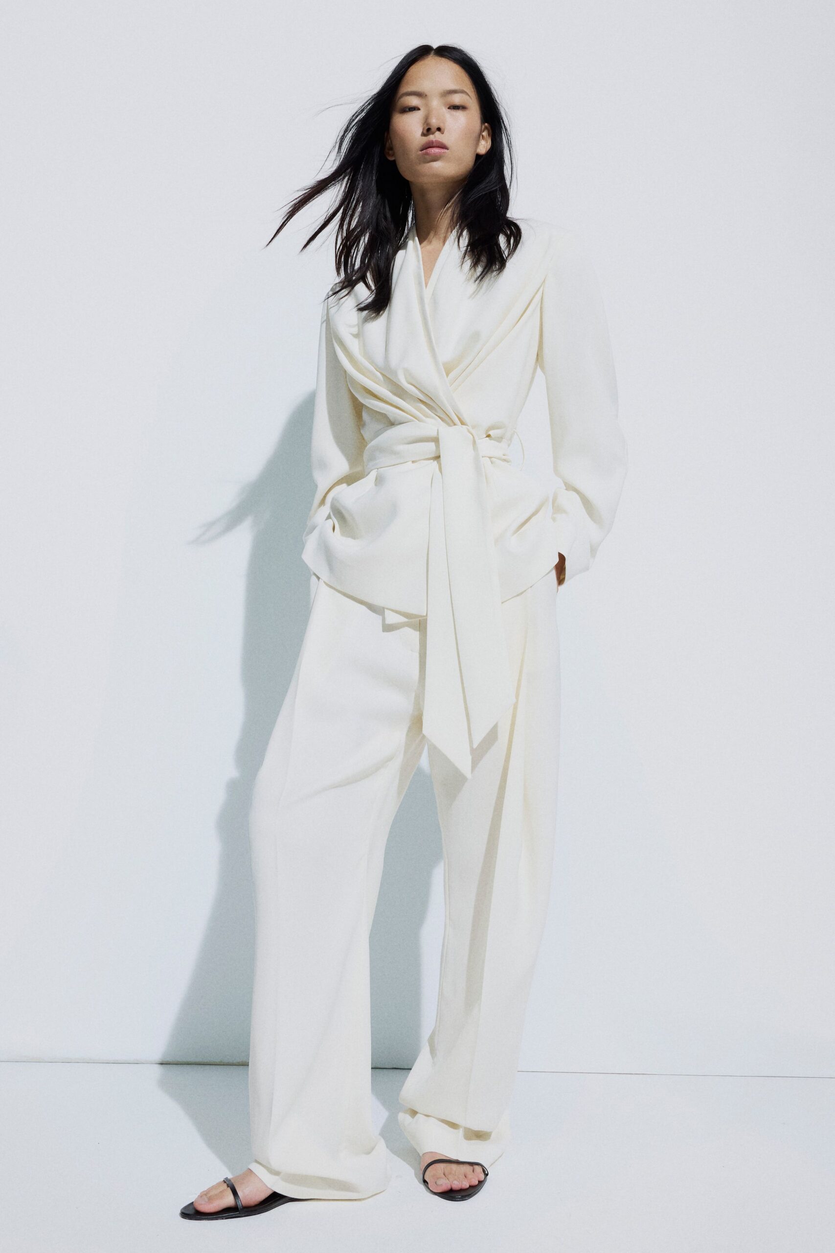Ivory crepe wrap blouse, deep shawl lapels and a long self-tie hanging low. Ivory wide-leg trousers, pressed clean and pooling over the instep. Black thong sandals, wafer-thin sole. First light in a hotel suite before the courthouse opens.