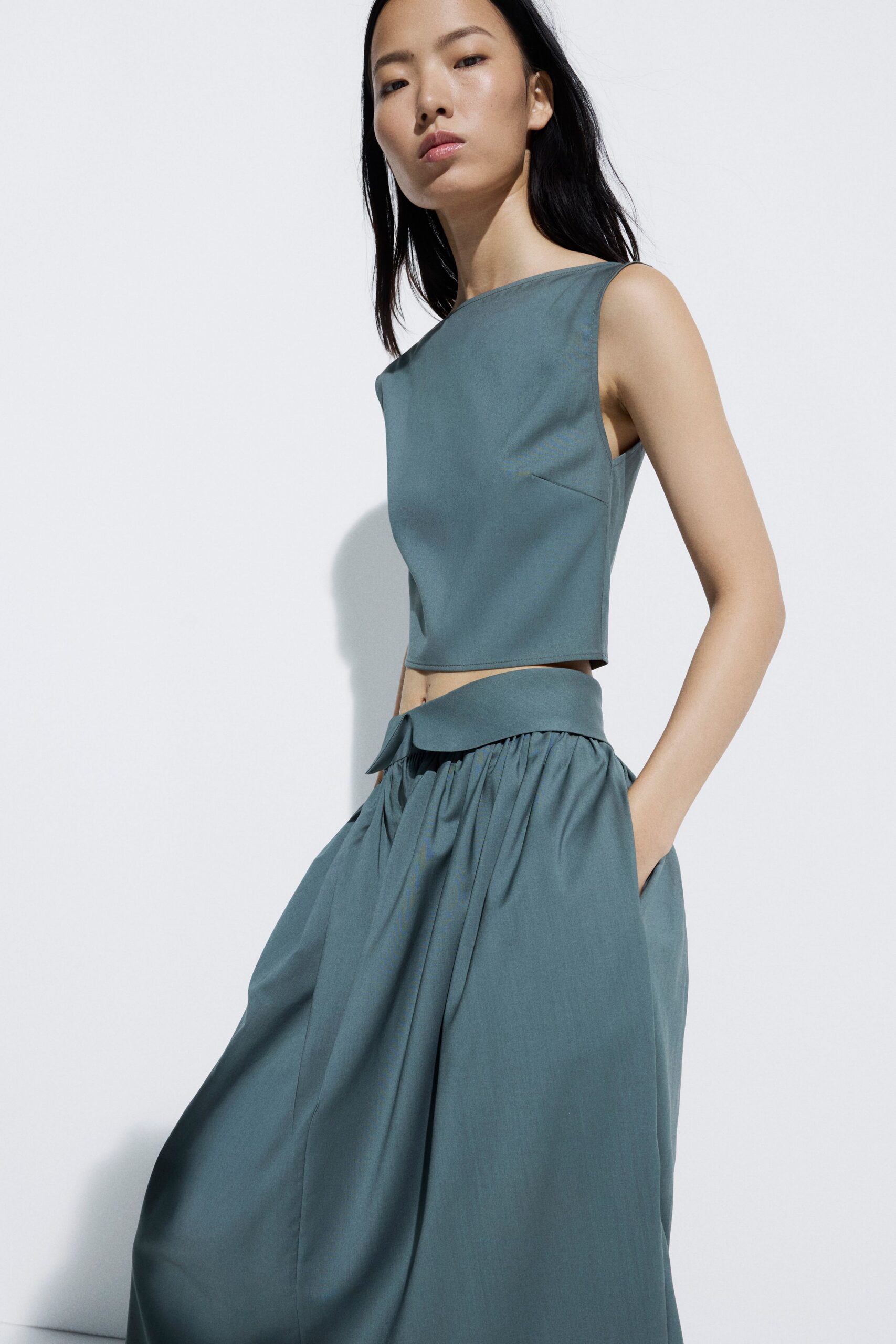 Dusty teal shell top with a clean bateau neckline and cropped hem. Matching wide trousers, softly gathered under a folded waistband tab. Hot afternoon gallery opening, concrete floors still holding the day's warmth.