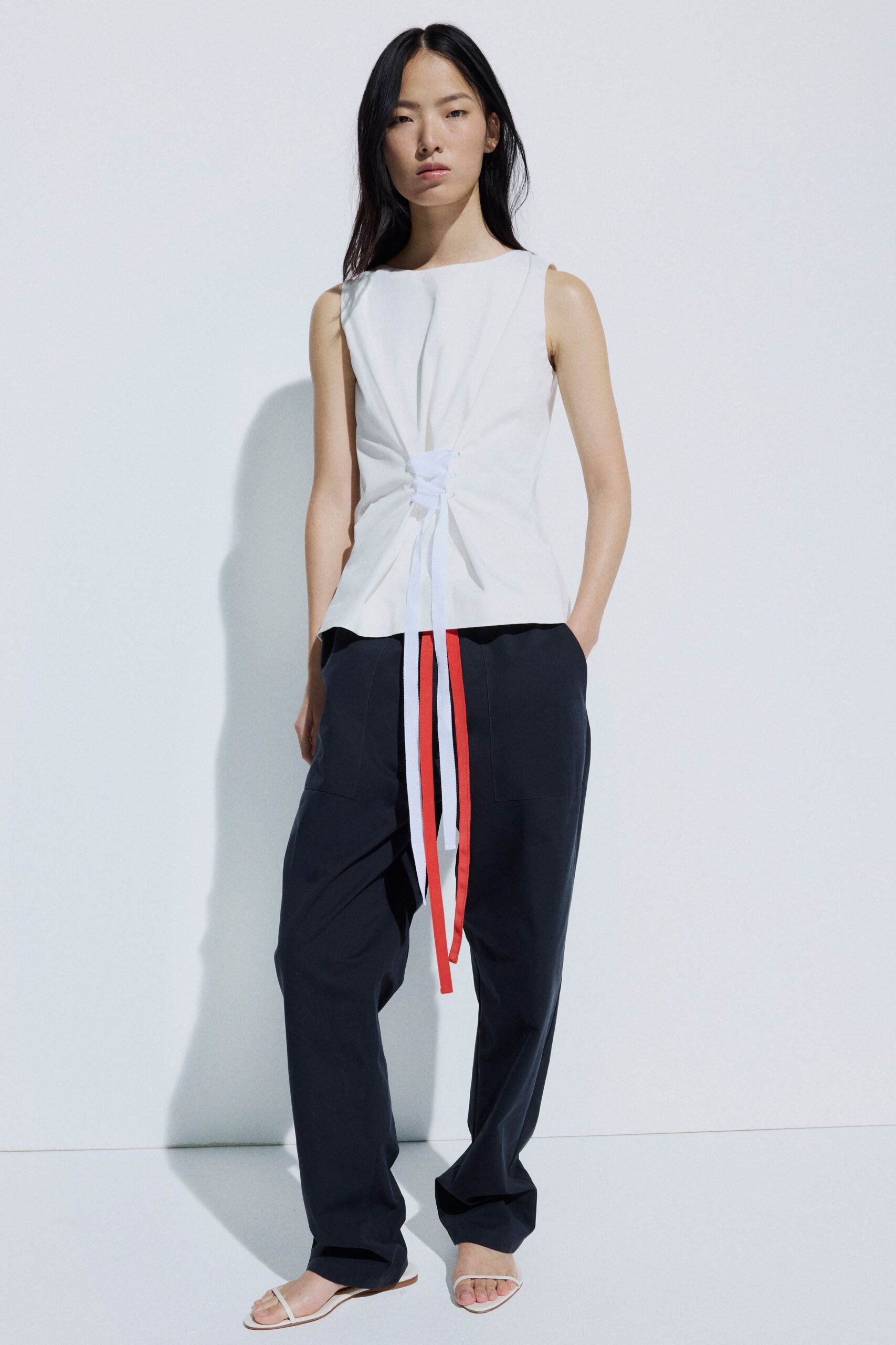 White sleeveless shell, waist cinched through a knotted slit with long ties. Ink straight trousers with oversized patch pockets and coral side stripes. Flat white thong sandals. Walking back from a museum courtyard into the heat off the pavement.