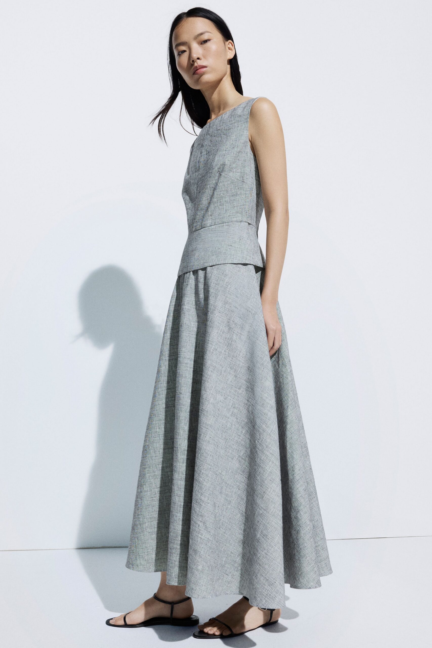 Pale grey slub-weave dress with a bateau neck and dropped peplum waist. Bare black thong sandals with narrow ankle straps. Museum courtyard at noon, heat bouncing off the stone.