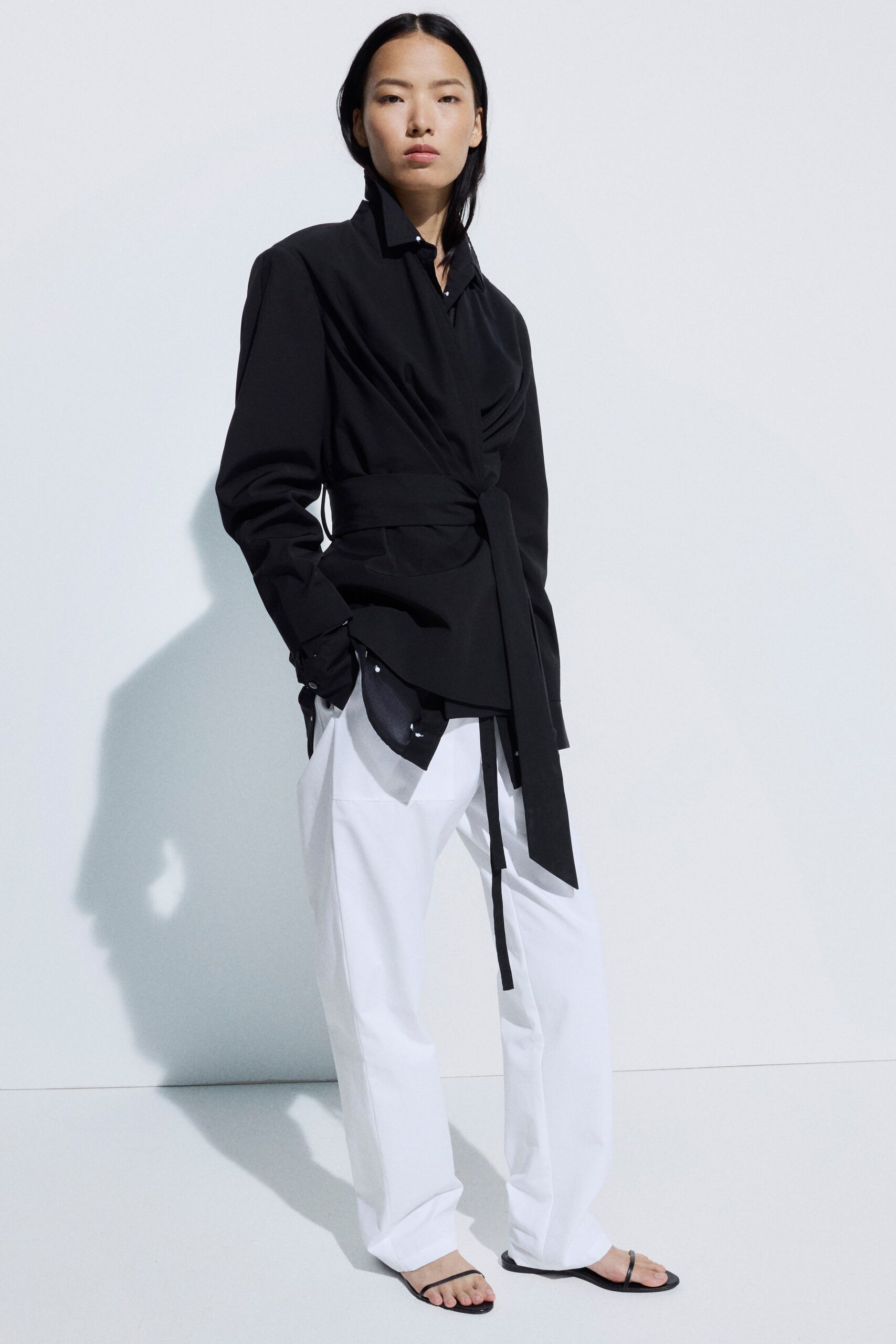 Black wrap shirt in crisp suiting cloth, ruched across one side. Wide self-belt knotted low, one long end hanging past the hip. White wide-leg trousers with pressed creases and pooled hems. Flat black thong sandals with barely there straps. Early ferry crossing, salt on the rail and paperwork in your bag.