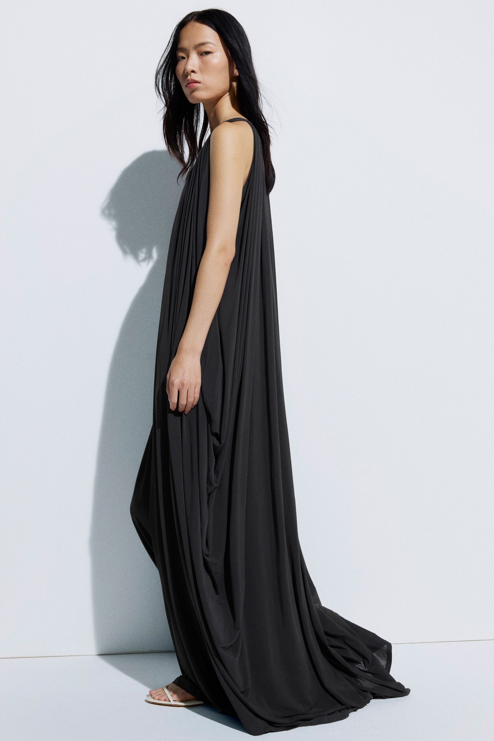 Black jersey column gown, knife pleats falling from thin shoulder straps into pooled side drapes. Barely there white thong sandals. Crossing a whitewashed terrace just before the heat lifts.