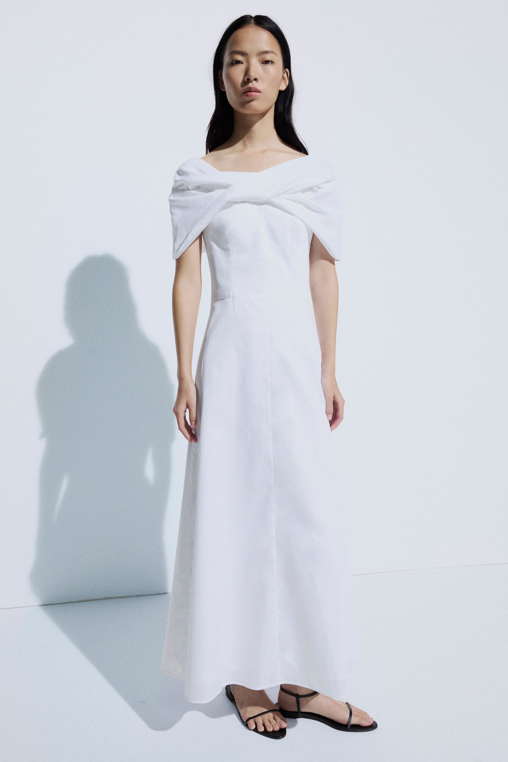 Ivory column dress with a folded off-shoulder band crossing into a soft knot. Black thong sandals with slender straps and a flat sole. Registry office at noon, carrying your shoes home in a paper bag.