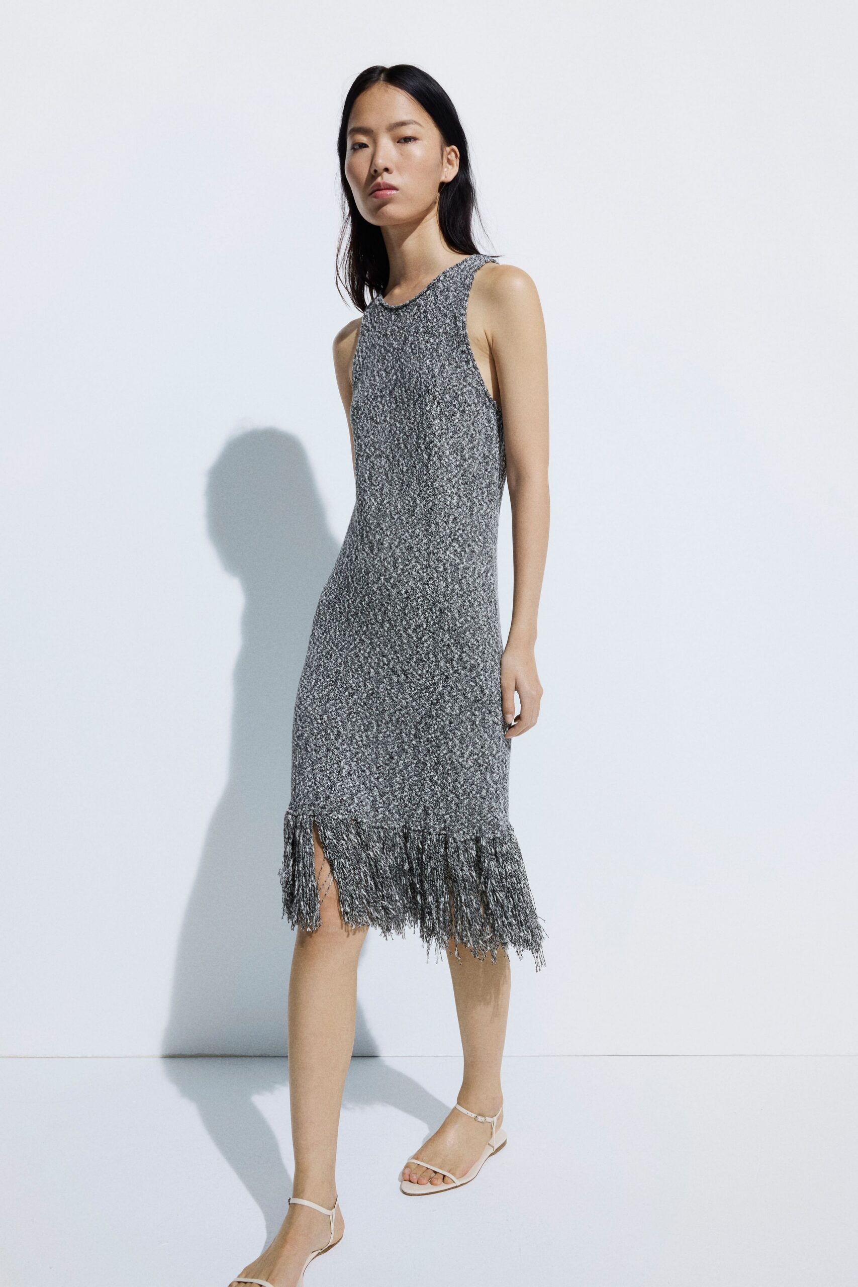 Marled grey knit tank dress, rib hugging close through the hips. Long cut fringe swinging from a dropped seam, one slit breaking it. Bare white ankle-strap sandals. Walking across a hotel terrace before the pool crowd comes up.