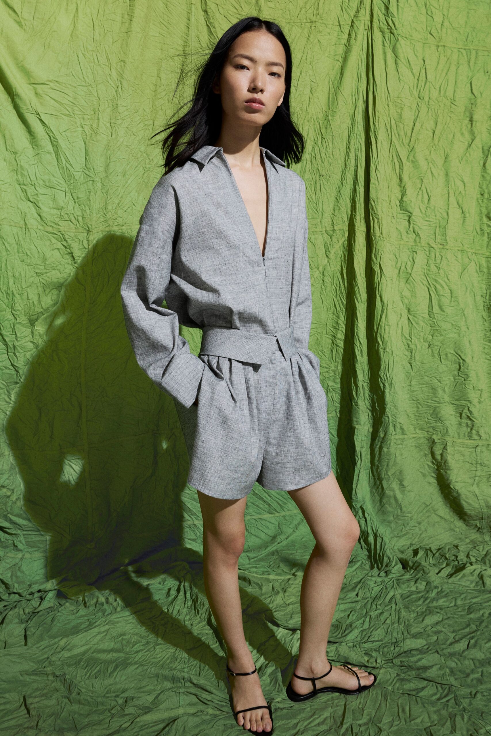 Pale grey marl romper with a deep split neck and dropped shoulders. Wide matching sash, knotted slightly off center at the waist. Black barely-there straps circling the ankle and toe. Walking back from a rental house pool before the towels dry.