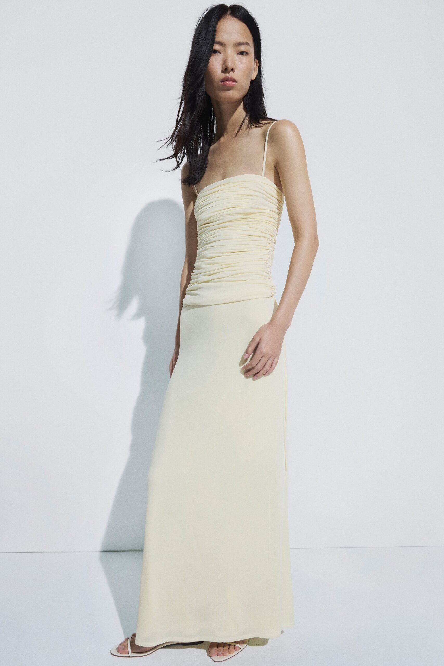 Buttercream column dress with ruched jersey bodice and thread-thin straps. Barely there white sandals, squared sole peeking past the hem. Morning after a beach wedding, finding coffee before anyone else wakes.