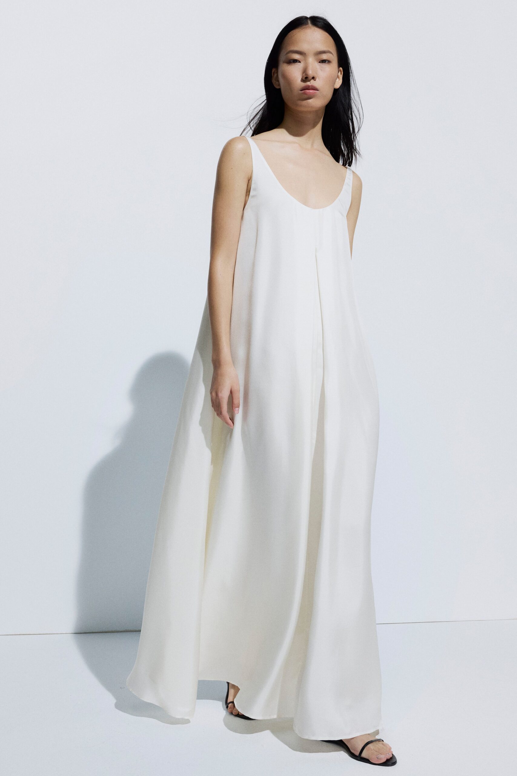 Ivory floor-length slip dress in fluid crepe, center pleat opening as she walks. Black thong sandals with narrow straps and flat soles. Crossing a whitewashed courtyard just after the stone has given up its heat.