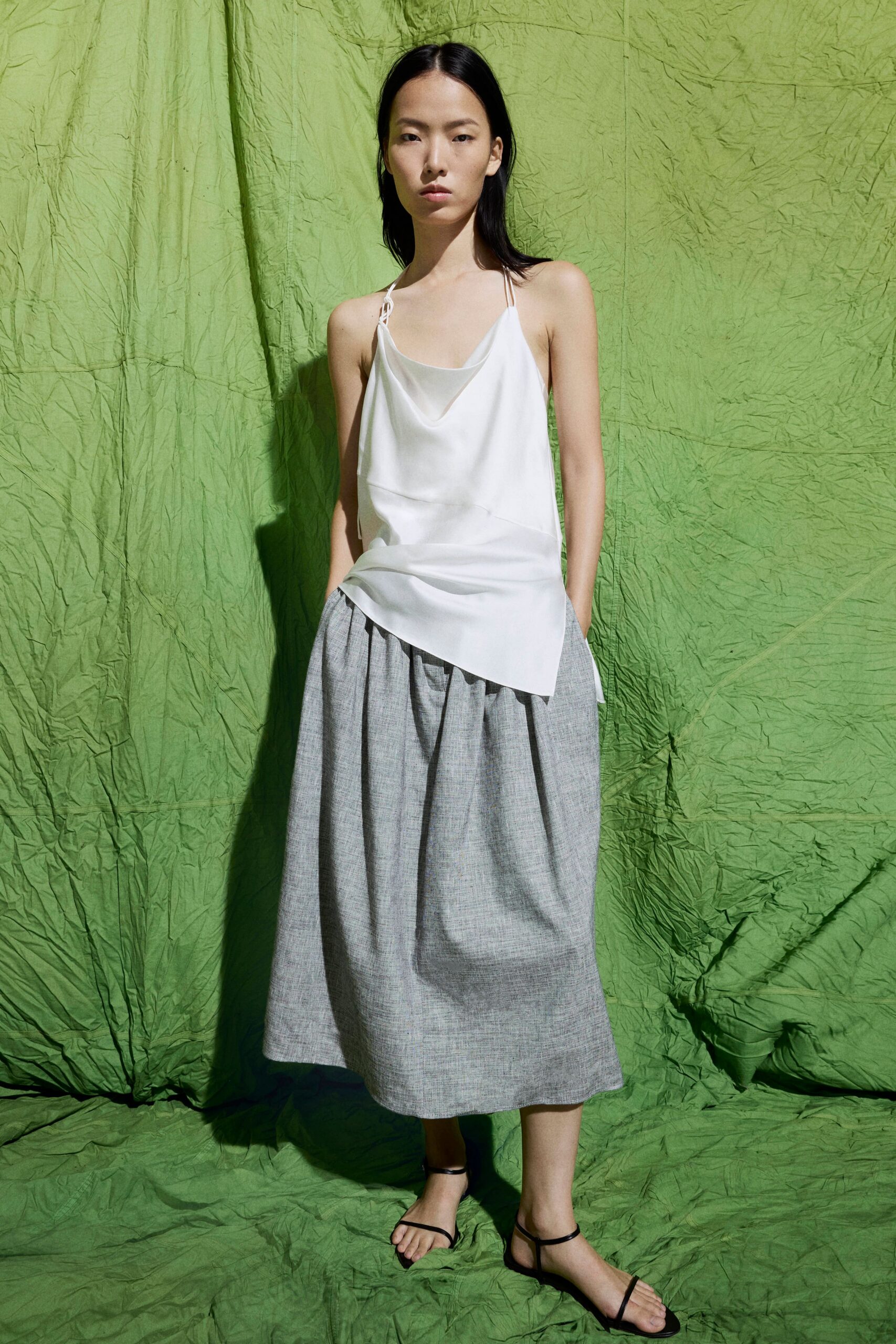 White satin camisole with a draped cowl and uneven handkerchief hem. Marl grey midi skirt, softly gathered into a full barrel shape. Black thong sandals with narrow ankle straps. Early ferry crossing, sea wind flattening the slip against your ribs.