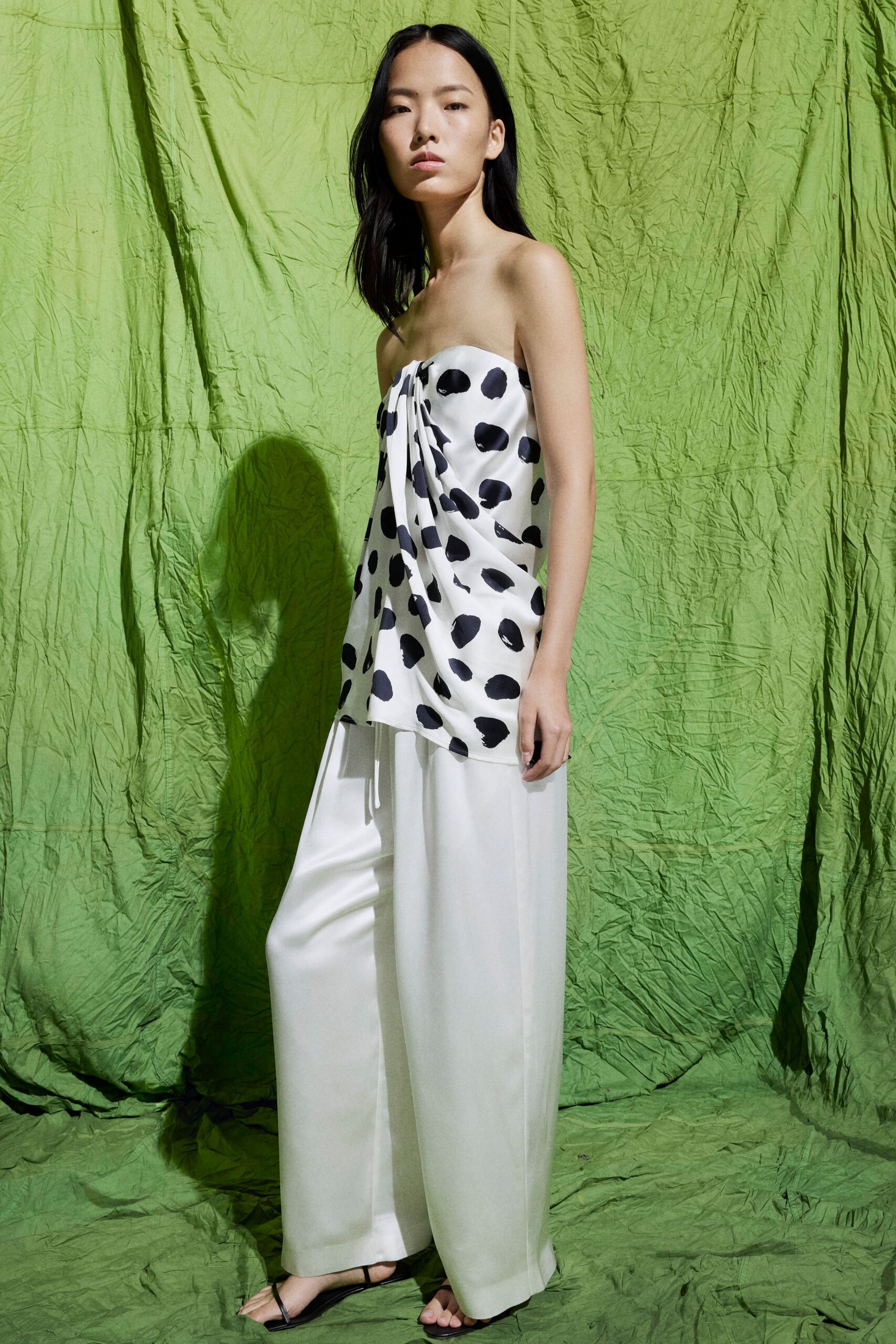 Strapless ivory satin top, brushstroke polka dots falling into a knotted drape. Fluid ivory trousers with a pressed front crease and full break. Black thong sandals with a bare, narrow strap. Crossing a hotel lawn after a wedding, heels already in hand.