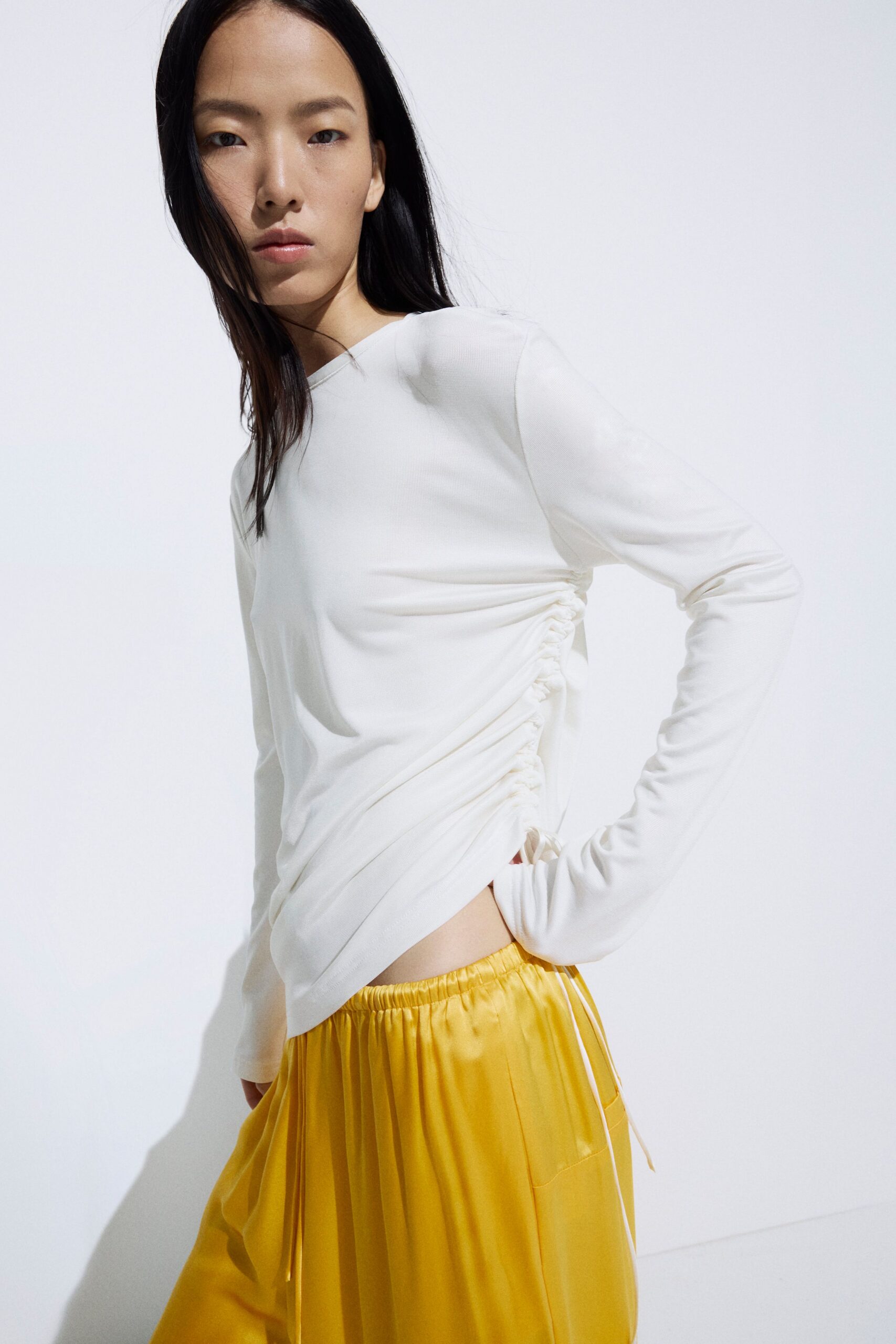 Ivory jersey top with side ruching and an angled hem exposing hip. Marigold satin trousers, softly pleated from a gathered elastic waist. Sun on the hallway wall, leaving the apartment before the heat sets in.