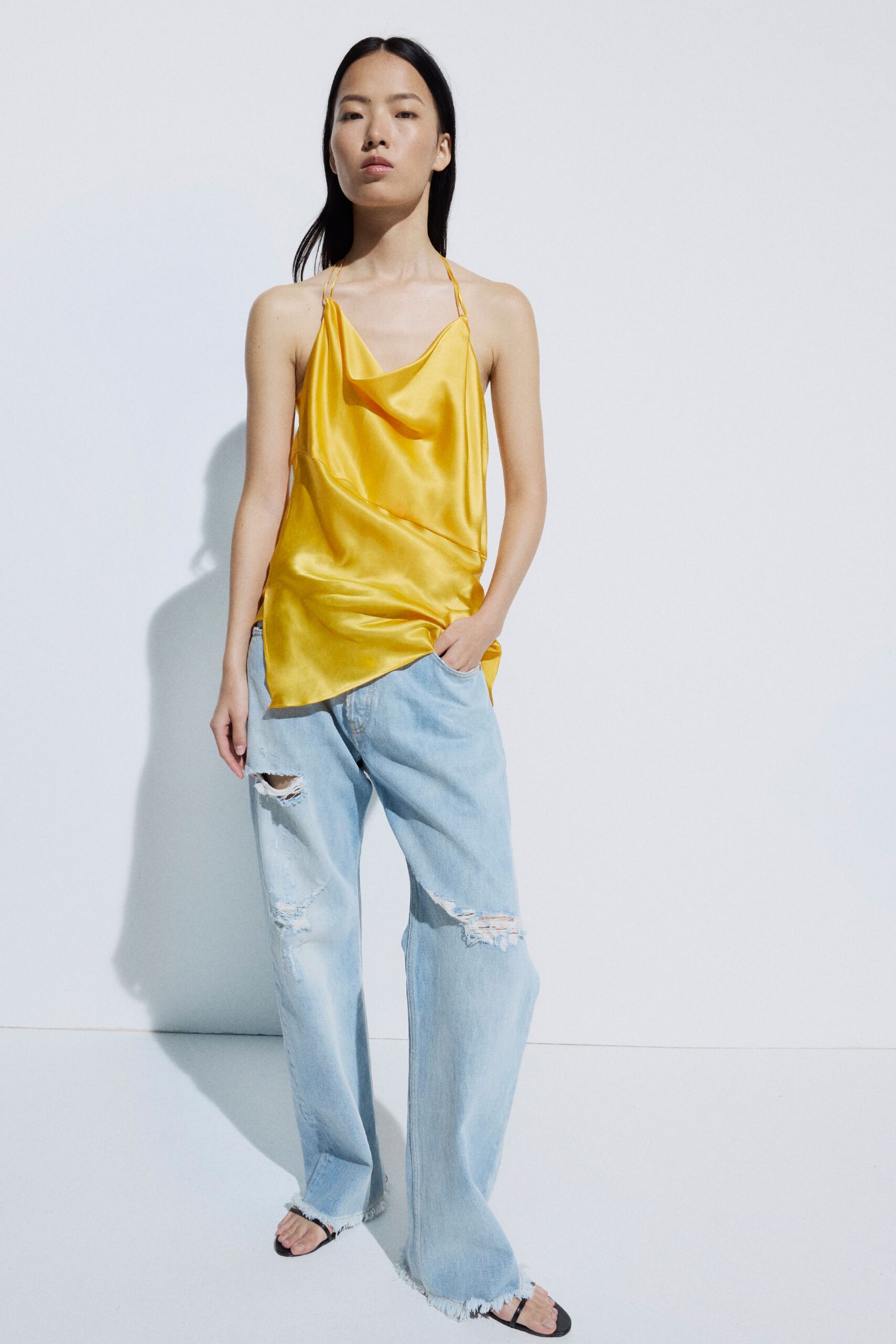 Butter-yellow satin camisole with a loose cowl and doubled spaghetti straps. Pale washed denim hanging wide and low, knees ripped to white threads. Black thong sandals, barely-there straps under frayed hems. Walking back from the beach bar after spilling mango juice down yourself.