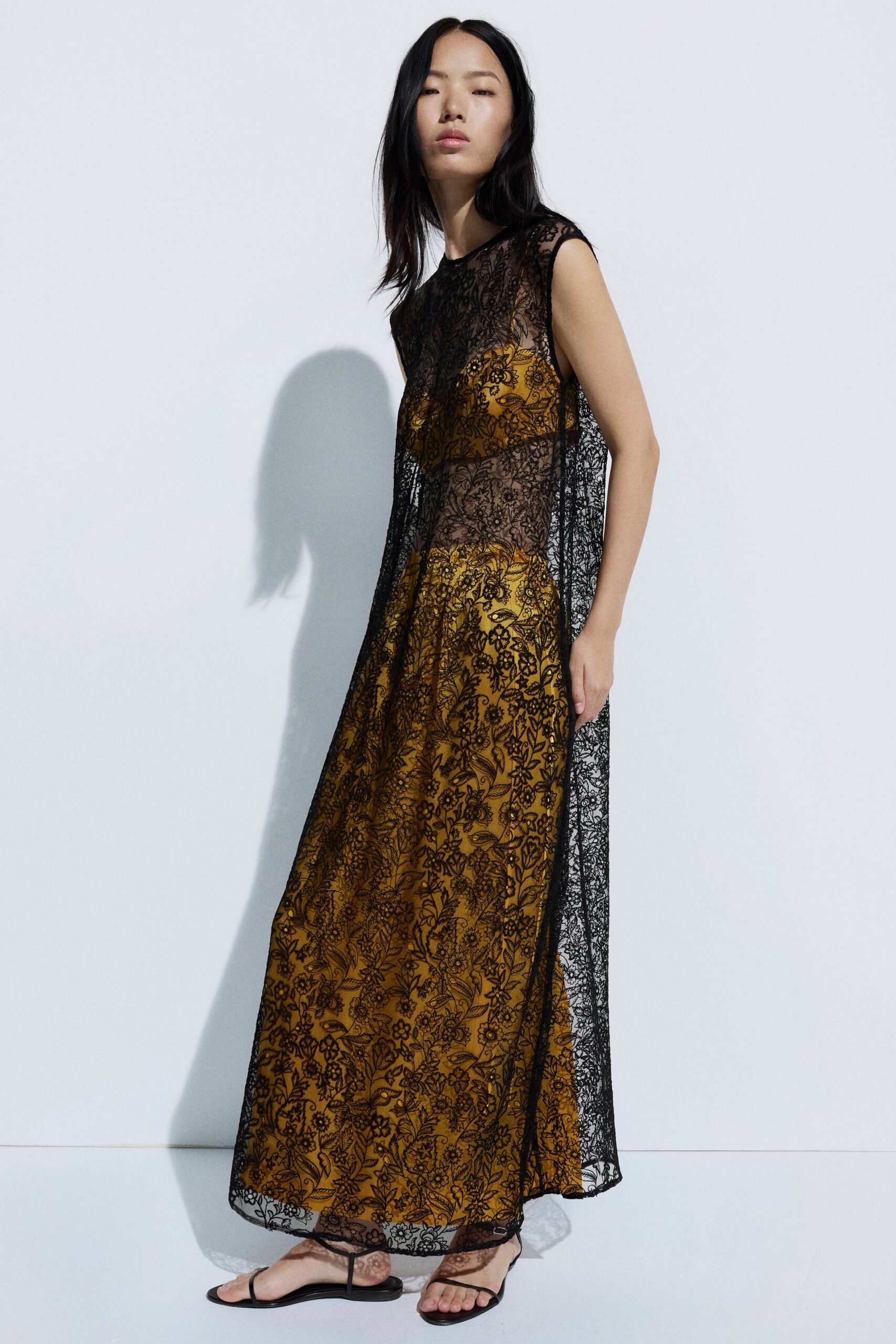 Saffron slip dress under a sheer black floral lace overdress, hem skimming the ankles. Black thong sandals with barely there straps. Afternoon wedding spillover, standing in the strip of shade by the church wall.