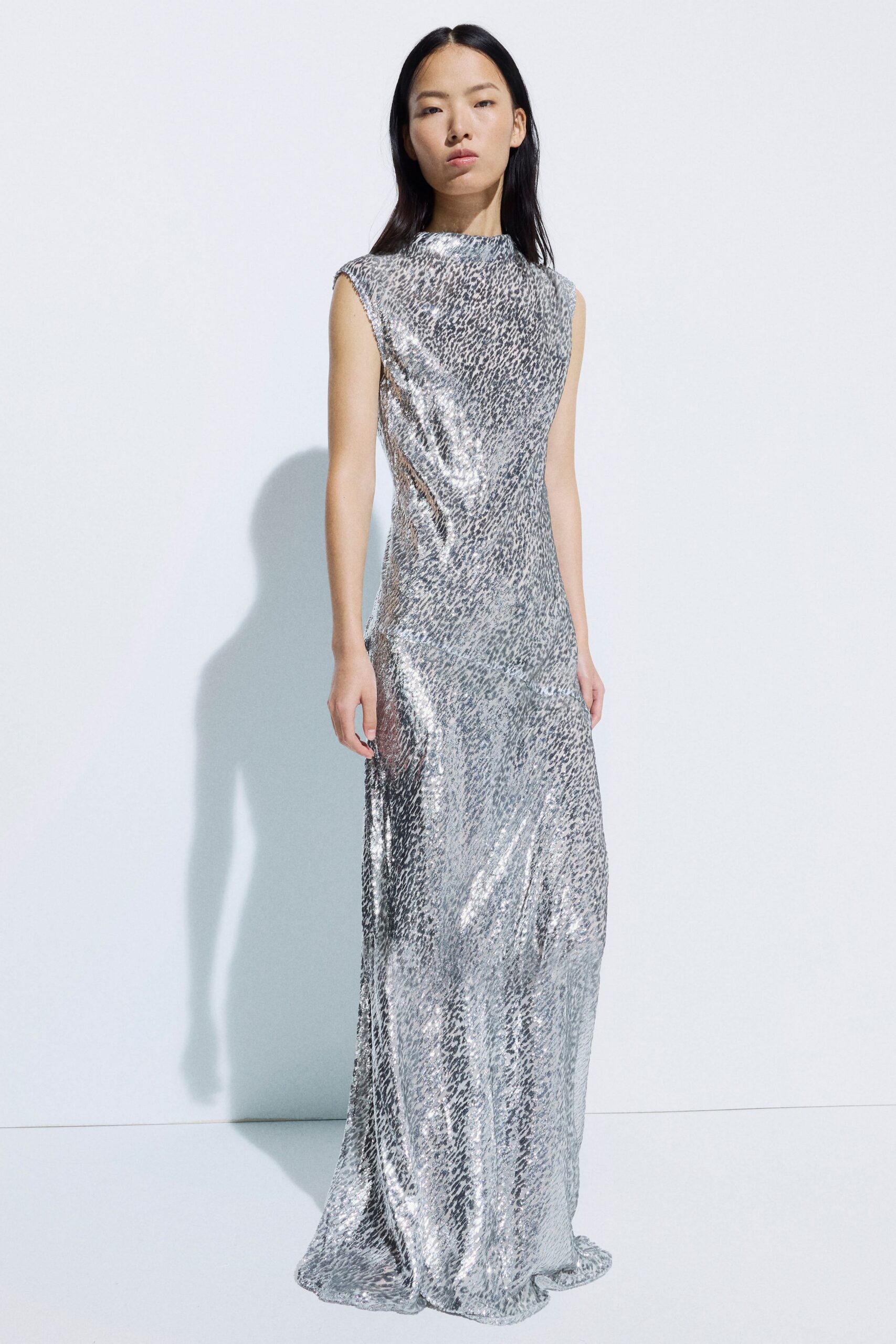 Silver leopard-sequined column gown, high neck and raw cap sleeves. Sheer mesh inset cutting up the left hip. Pooled floor-length hem with a narrow twist at center front. Whatever catches the light hardest at a hotel ballroom fundraiser.