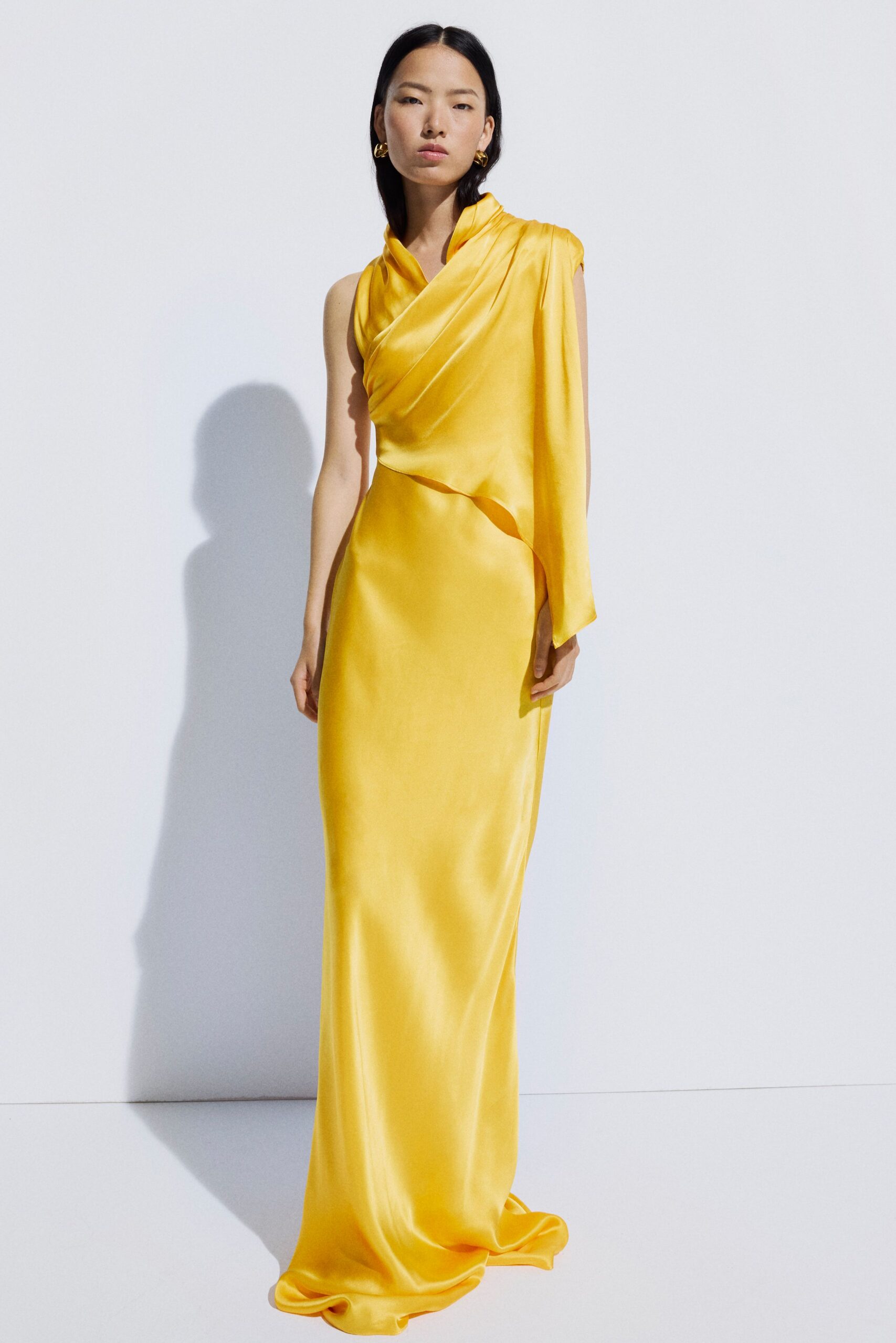 Canary satin column gown with a draped scarf panel falling from one shoulder. Small crumpled gold hoops. Sun hitting the stairwell outside a registry office at noon.