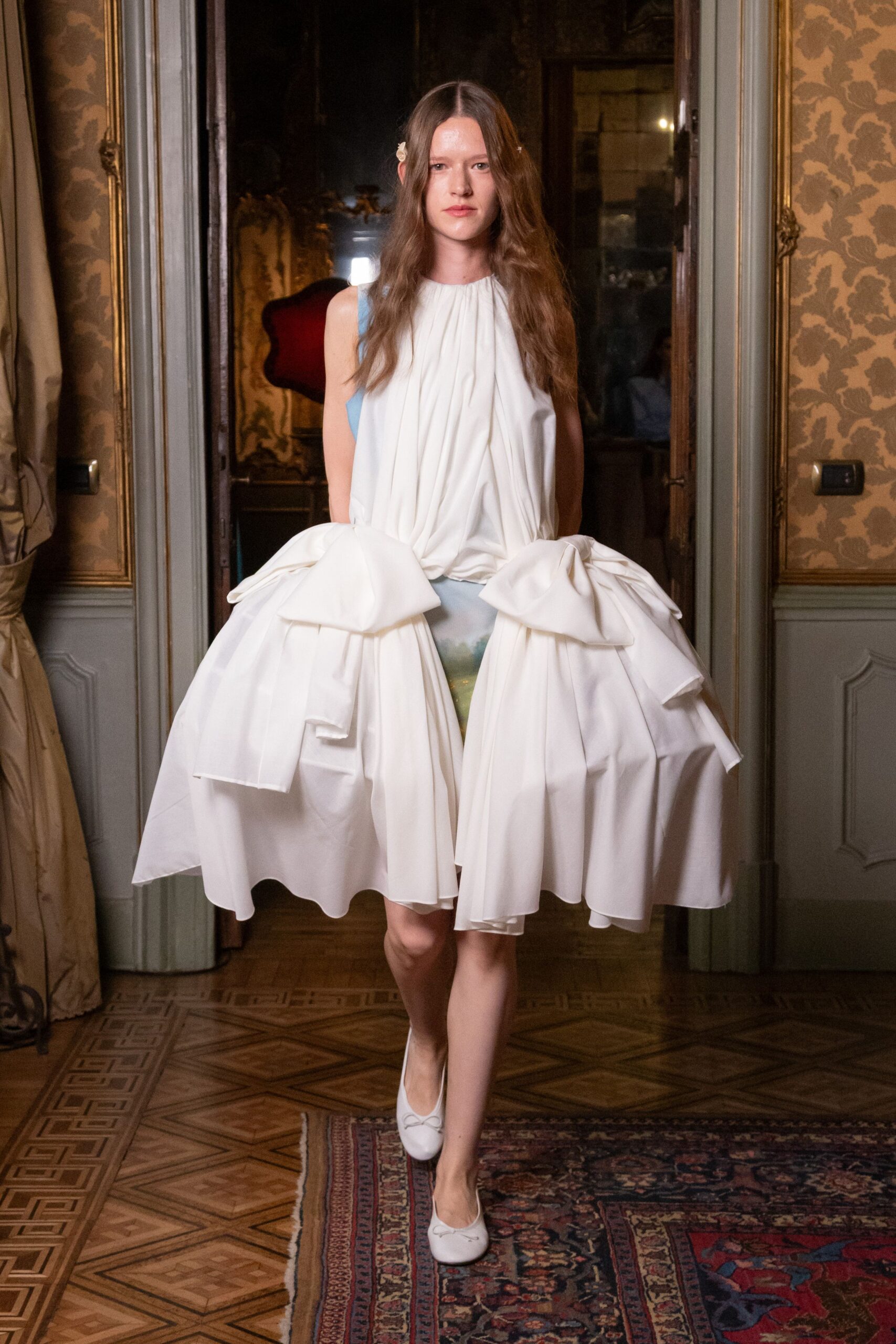 White satin dress with a gathered bodice and enormous folded pannier bows. White ballet pumps with tiny bow trims at the vamp. Small white flower clips pinned beside the center part. Crossing a parquet salon after the cake table has been cleared.