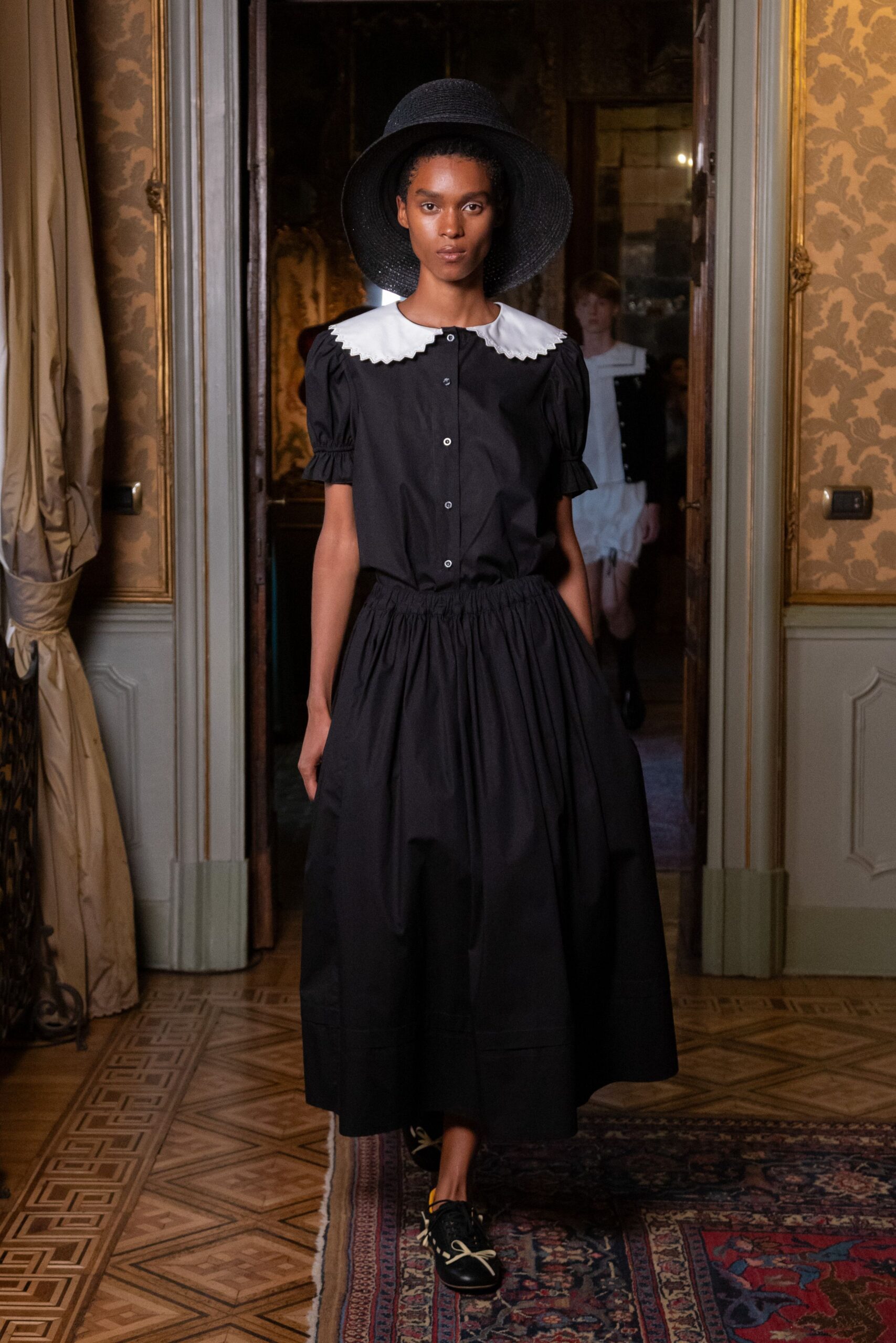 Wide-brim black straw hat with a tightly woven crown. Black cotton blouse with a white scalloped collar and puff sleeves. Full black poplin skirt, densely gathered into an elastic waist. Black leather lace-ups with oversized cream bow laces. Crossing the parquet for lunch in a faded villa with the shutters open.