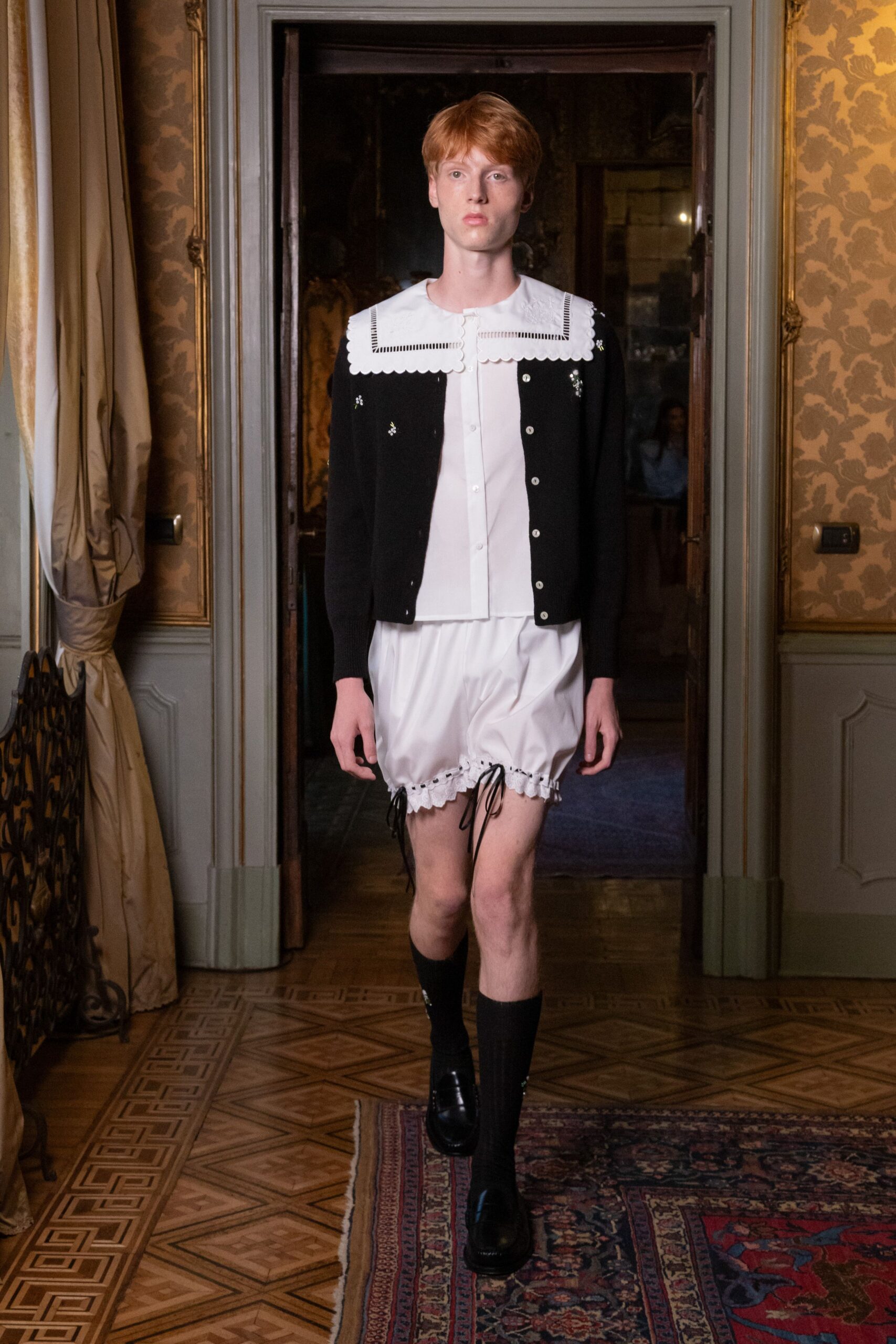 Black knit cardigan with tiny floral beading and white buttons. White blouse topped by an oversized scalloped bib collar. White bloomers gathered at the thigh with black tie cords. Black ribbed knee socks. Glossy black loafers with thick soles. Crossing creaky parquet to answer the door in a rented villa at dusk.