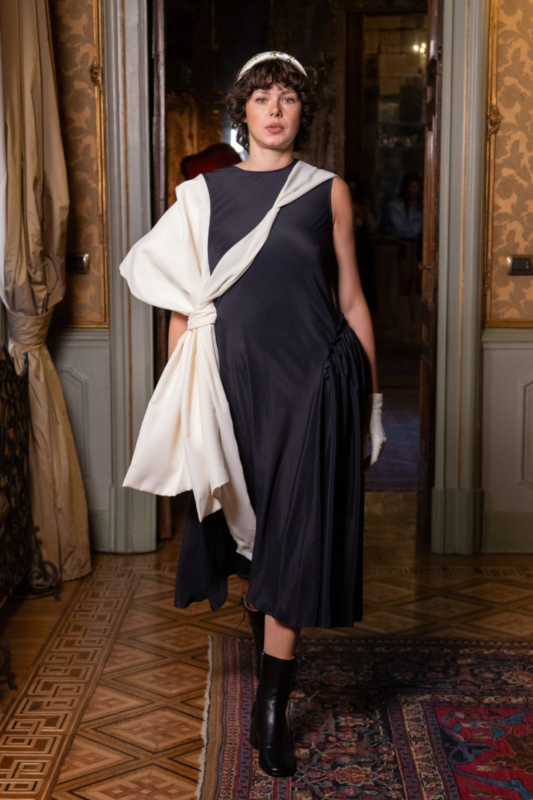 White scarf tied close over the crown. Ink jersey dress with a cream duchesse panel knotted across the torso. Single white glove. Black leather ankle boots, square at the toe. Crossing a grand hotel corridor after slipping out of the wedding dinner.