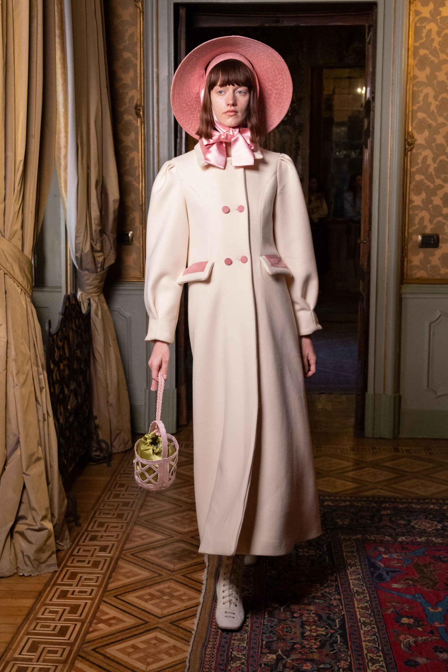 Wide pink straw hat tied under the chin with satin bows. Ivory floor-length coat with puffed sleeves, pink-covered buttons and pocket flaps. Small lattice bucket bag with a twisted cord handle and chartreuse lining. White lace-up boots peeking from the hem. Sunday christening lunch in a drafty villa, gravel still on the soles.