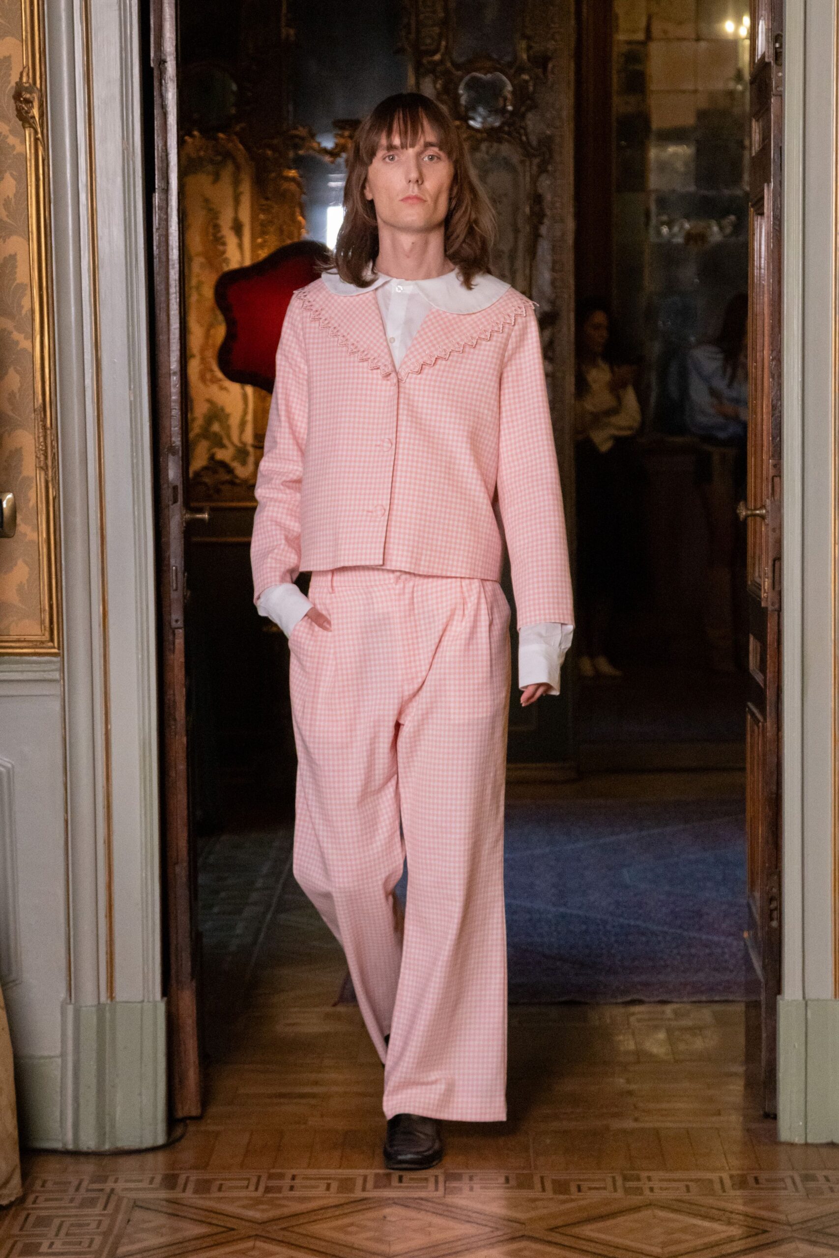 White bib-front blouse with a rounded collar and long cuffs. Pink gingham jacket edged with zigzag trim, fastening on one visible button. Matching gingham trousers cut loose through the leg. Dark square-toe shoes. Crossing a faded palazzo salon before anyone has opened the shutters.