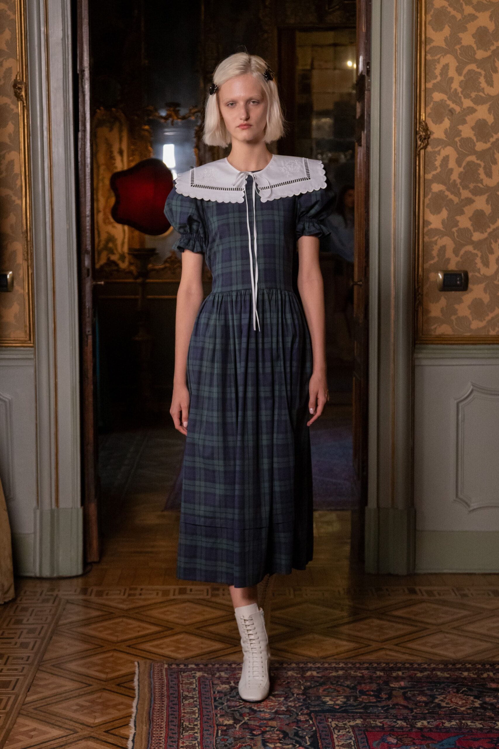 Black patent barrettes pinning the blunt bob at both temples. Blue-green tartan dress with puff sleeves and a scalloped white bib collar. White lace-up ankle boots with a neat row of punched eyelets. Crossing a drafty manor hallway before anyone rings for tea.