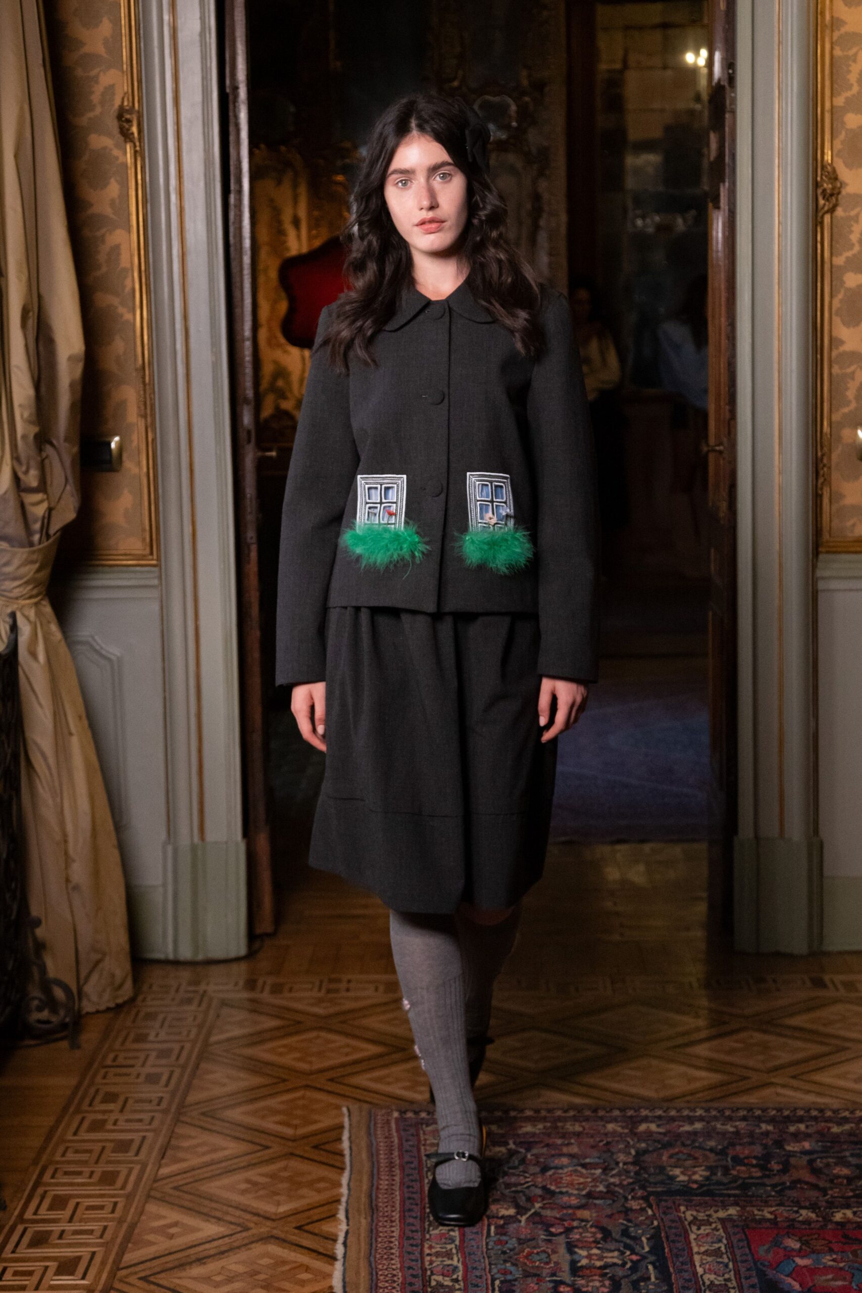 Black flower clip tucked above one ear. Charcoal wool jacket with rounded collar, appliqued windows and green feather trim. Matching charcoal skirt cut full below the knee. Grey ribbed knee socks. Black Mary Janes with a small silver buckle. Crossing a draughty parlour as the house wakes up around you.