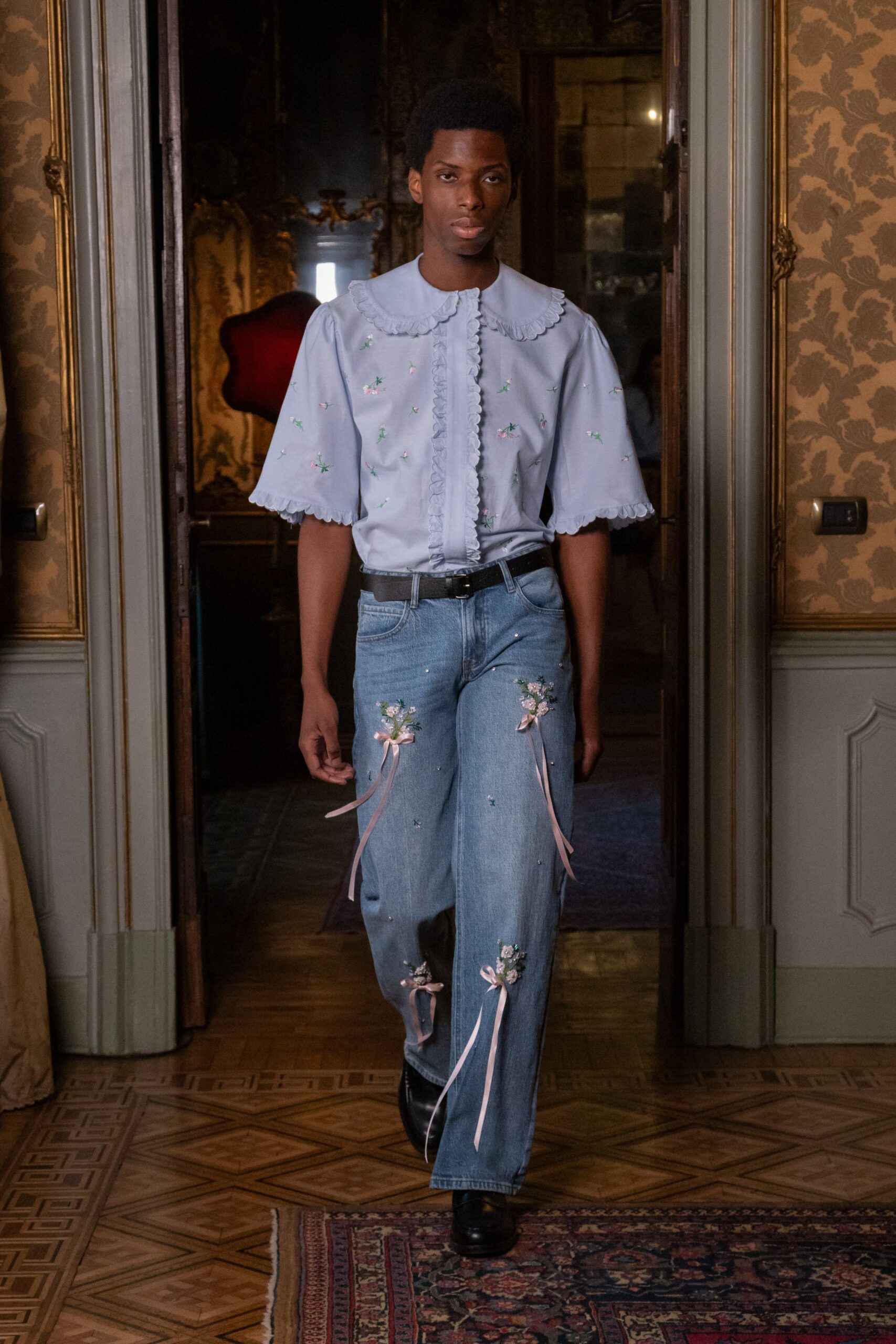 Powder-blue blouse with scalloped Peter Pan collar and ruffled placket. Black leather belt with a plain square buckle. Light-wash straight jeans embroidered with posies and trailing pink ribbon bows. Black leather boots, blunt at the toe. Crossing a faded palazzo salon with cake crumbs still on the plate.