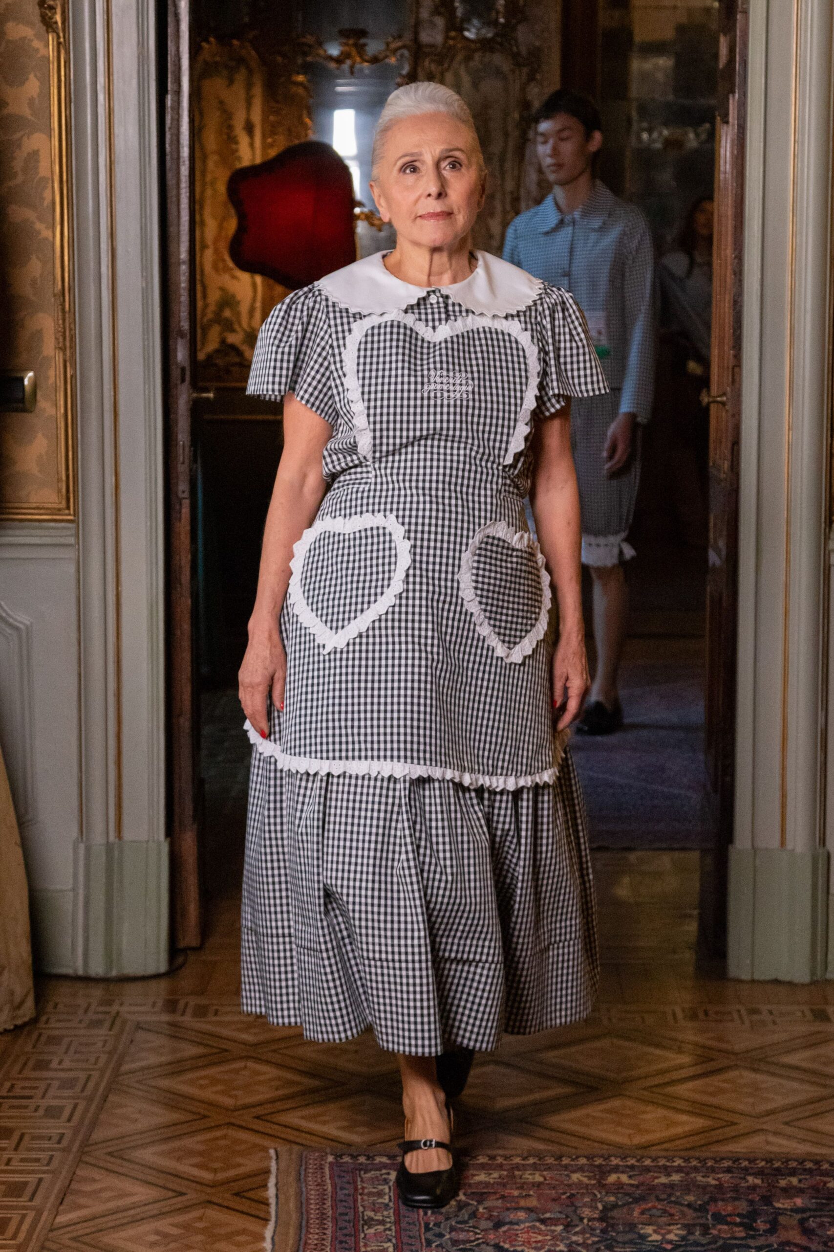 Black-and-white gingham dress with a broad scalloped white collar. Heart-shaped apron panels edged in white ricrac, monogram stitched at center. Black Mary Jane pumps with a tiny silver buckle. Crossing a faded palazzo salon with the good silver already laid out.