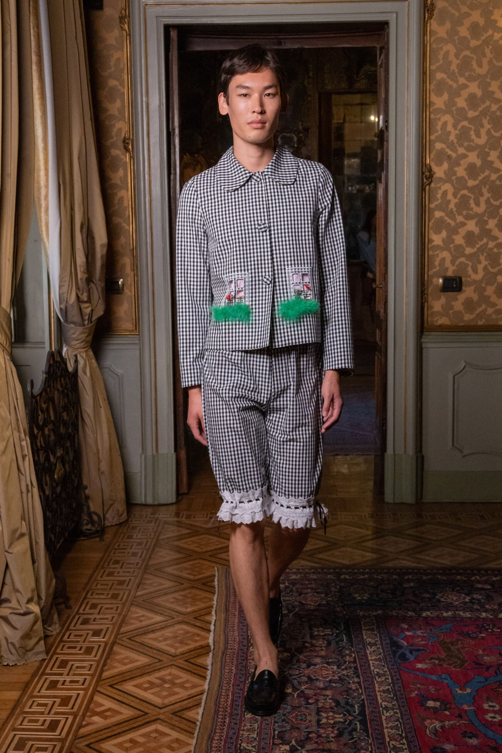 Black-and-white gingham jacket with a rounded collar and appliqued cottage windows. Matching knee shorts, hemmed in white eyelet ruffles with black ribbon lacing. Black leather loafers. Crossing a parquet salon dressed for tea in somebody else's country house.