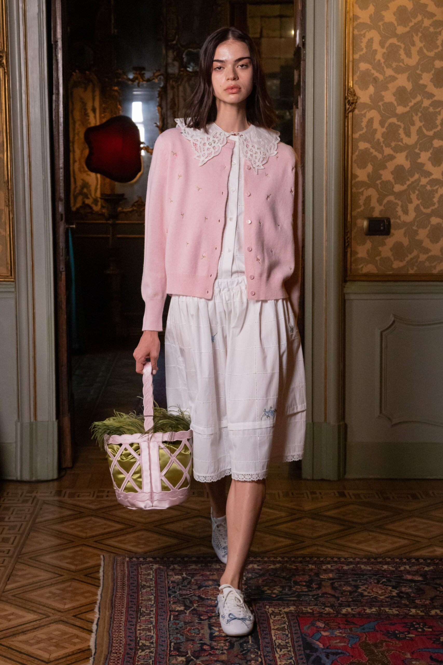 Powder-pink knit cardigan dotted with tiny stitched sprigs. White cotton dress with a broad cutwork lace collar and pintucked panels. Pink lattice bucket bag lined in lime satin, green feathers spilling out. White lace-up flats with pale blue floral embroidery. For carrying Easter grass through a drafty palazzo hallway.
