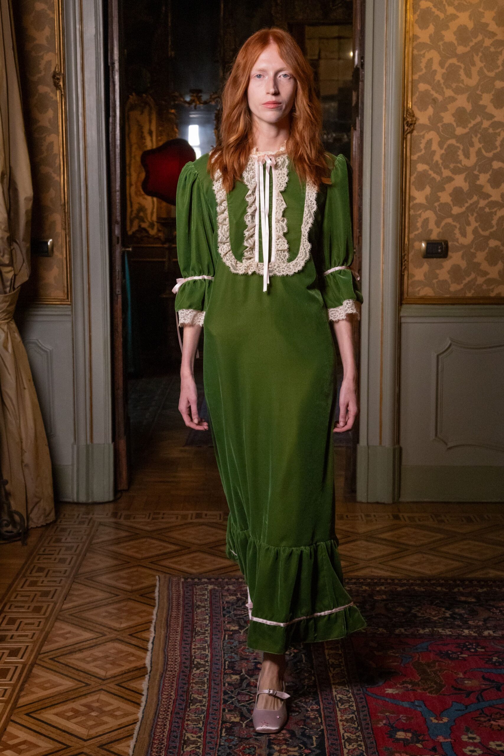 Moss velvet column dress with cream lace bib, puffed sleeves, pink ribbon ties. Powder-pink satin Mary Janes with a tiny embroidered flower at the toe. Crossing a draughty palazzo hallway before anyone else is awake.