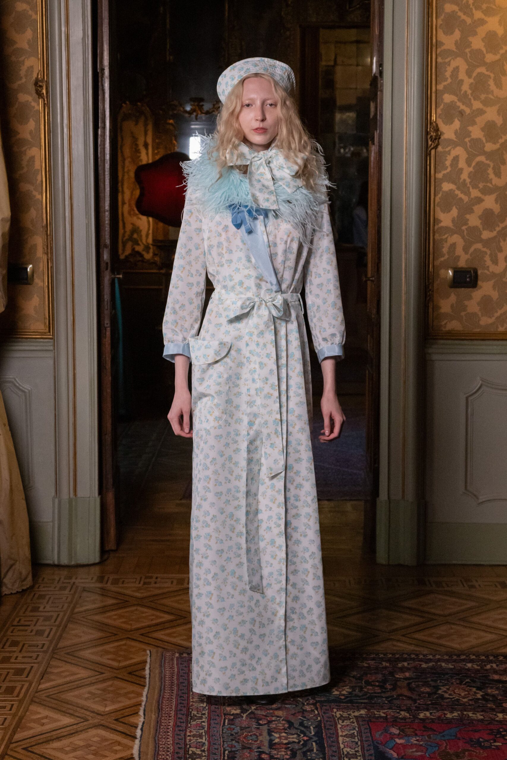 Quilted beret in the same faded blue sprig print. Oversized neck bow in crisp floral cotton. Floor-length wrap dress in powder-blue sprig cotton, one patch pocket low. Pale blue feather collar tied on with grosgrain ribbons. Morning in a drafty villa, carrying breakfast on a silver tray.