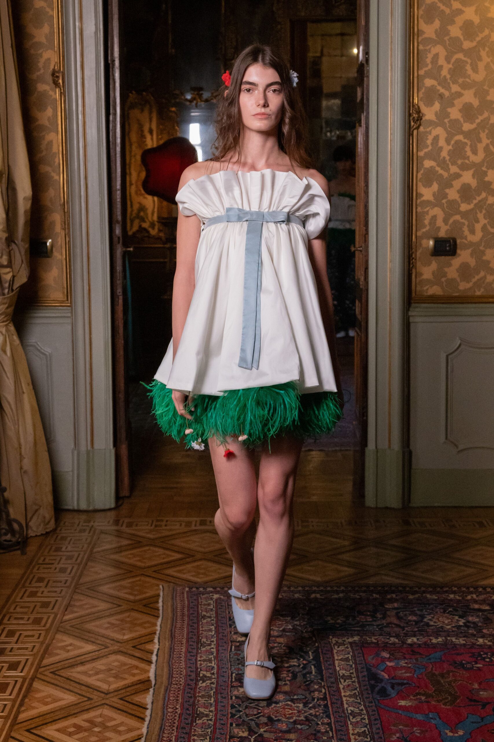Red and powder-blue flowers tucked behind the ears. Ivory satin babydoll dress, cartridge-pleated neckline bound with blue ribbon. Emerald ostrich feathers bursting from the mini hem. Powder-blue Mary Janes with slim silver buckles. Crossing a faded palazzo drawing room after midnight cake has been cut.