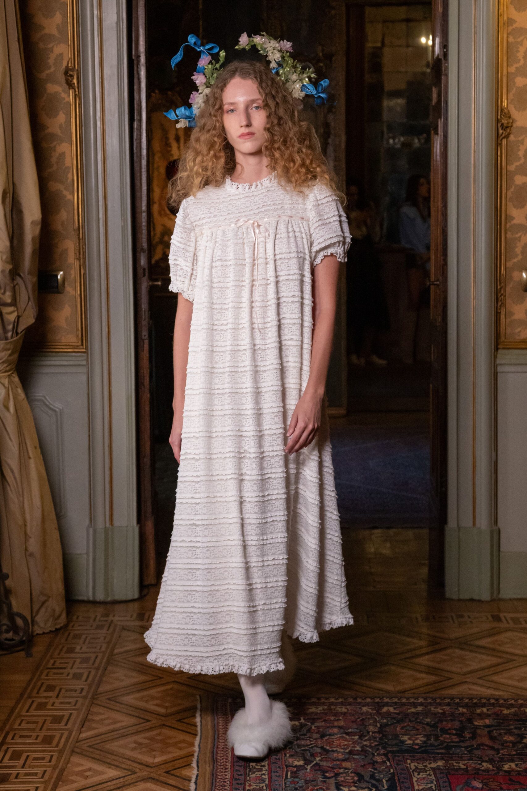 Loose ring of white blossoms tied with bright blue ribbon bows. Ivory ankle-length dress in horizontal lace bands, ribbon tie drooping from the yoke. Opaque white tights. White flats buried under fluffy marabou. Crossing cold parquet at dawn after sleeping in a borrowed manor.