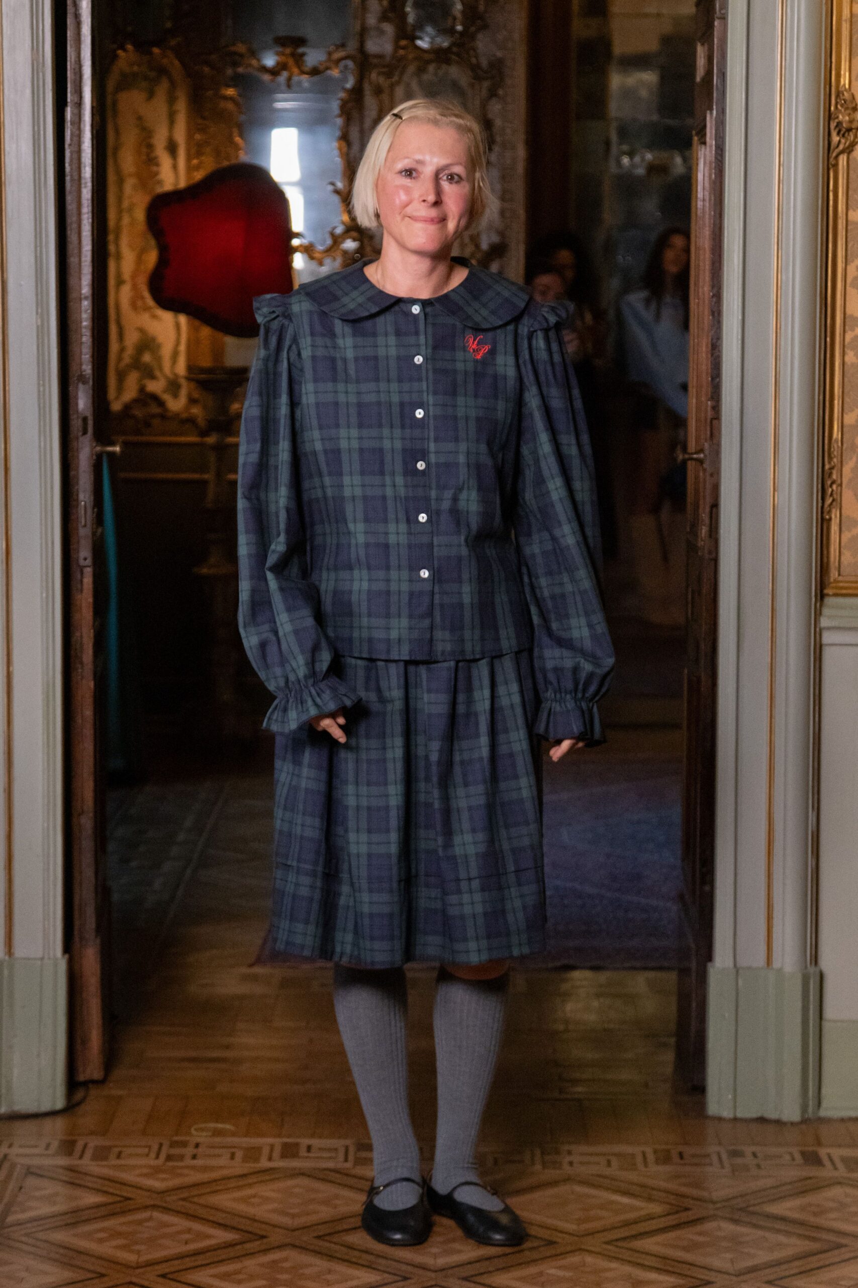Blue-green tartan blouse with a broad Peter Pan collar and puffed sleeves. Matching tartan skirt, softly gathered into a dropped waist seam. Grey ribbed knee socks. Black Mary Jane flats with rounded toes. Crossing a draughty manor hallway before tea is laid out.