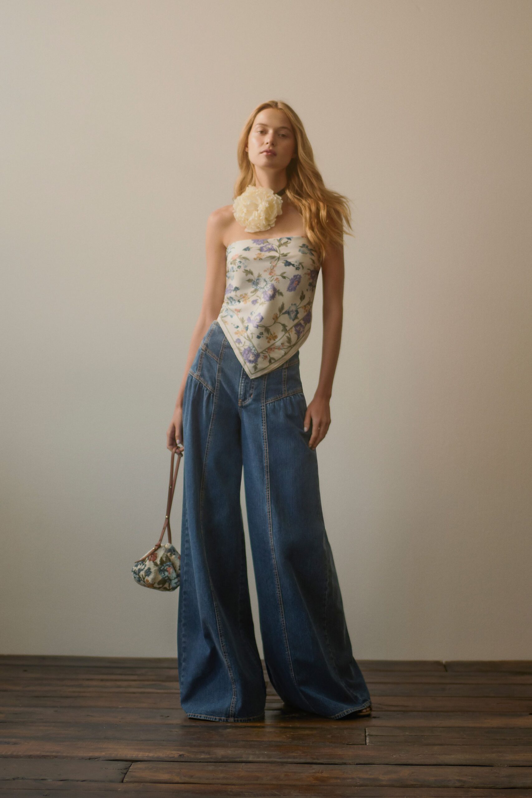 Oversized cream rosette at the throat on a narrow dark band. Silky ivory floral scarf top, pointed hem draped over the waistband. Dark indigo wide-leg jeans with curved panel seams and deep volume. Tiny floral pouch with a thin brown wrist strap. Sunday house viewing, still carrying cut flowers from the market.