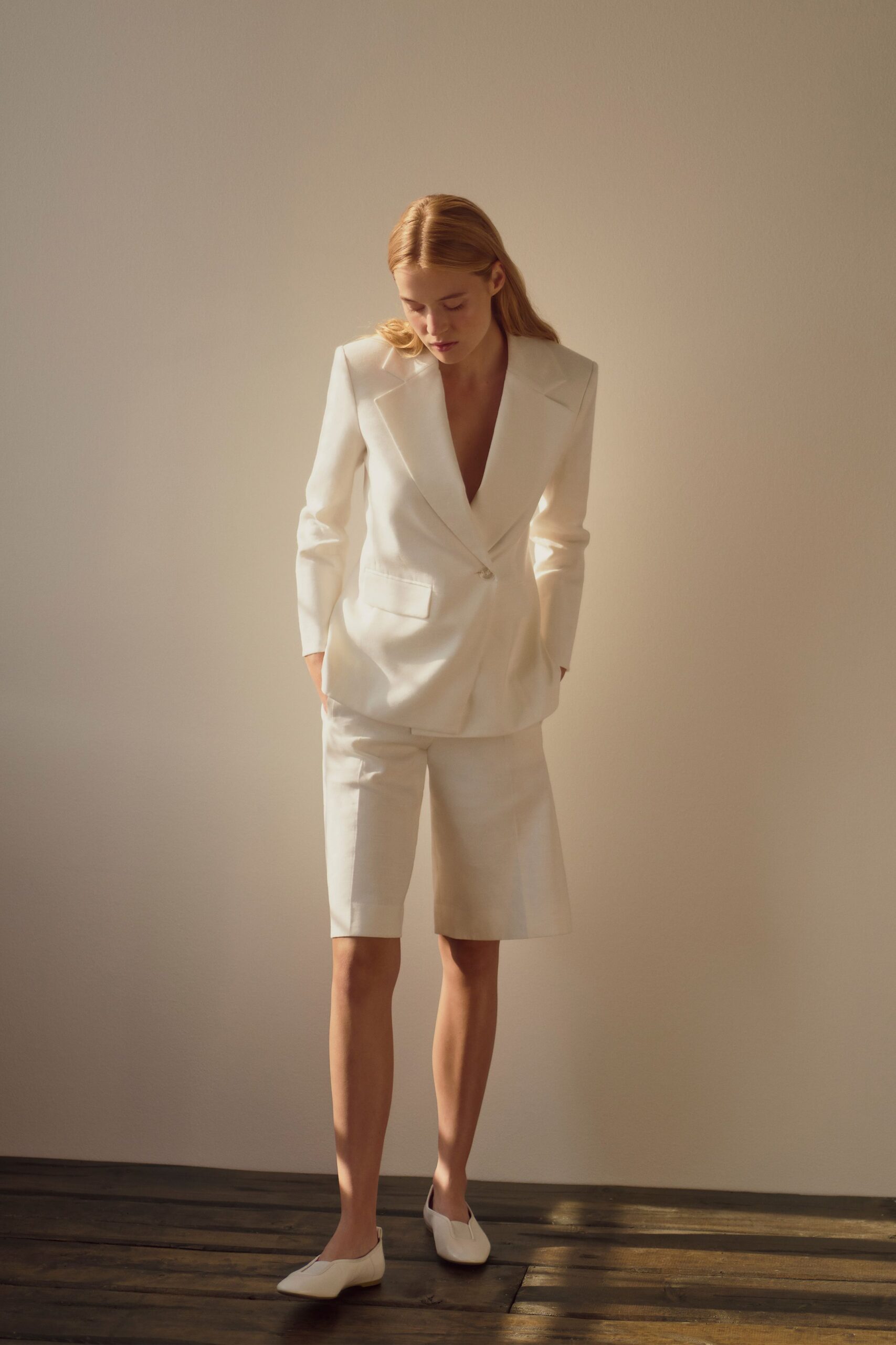 Ivory suiting blazer with sharp padded shoulders and a single low button. Matching knee-length tailored shorts with a clean pressed front. White leather flats with a narrow split at the vamp. Morning light in a rented room before the civil ceremony.