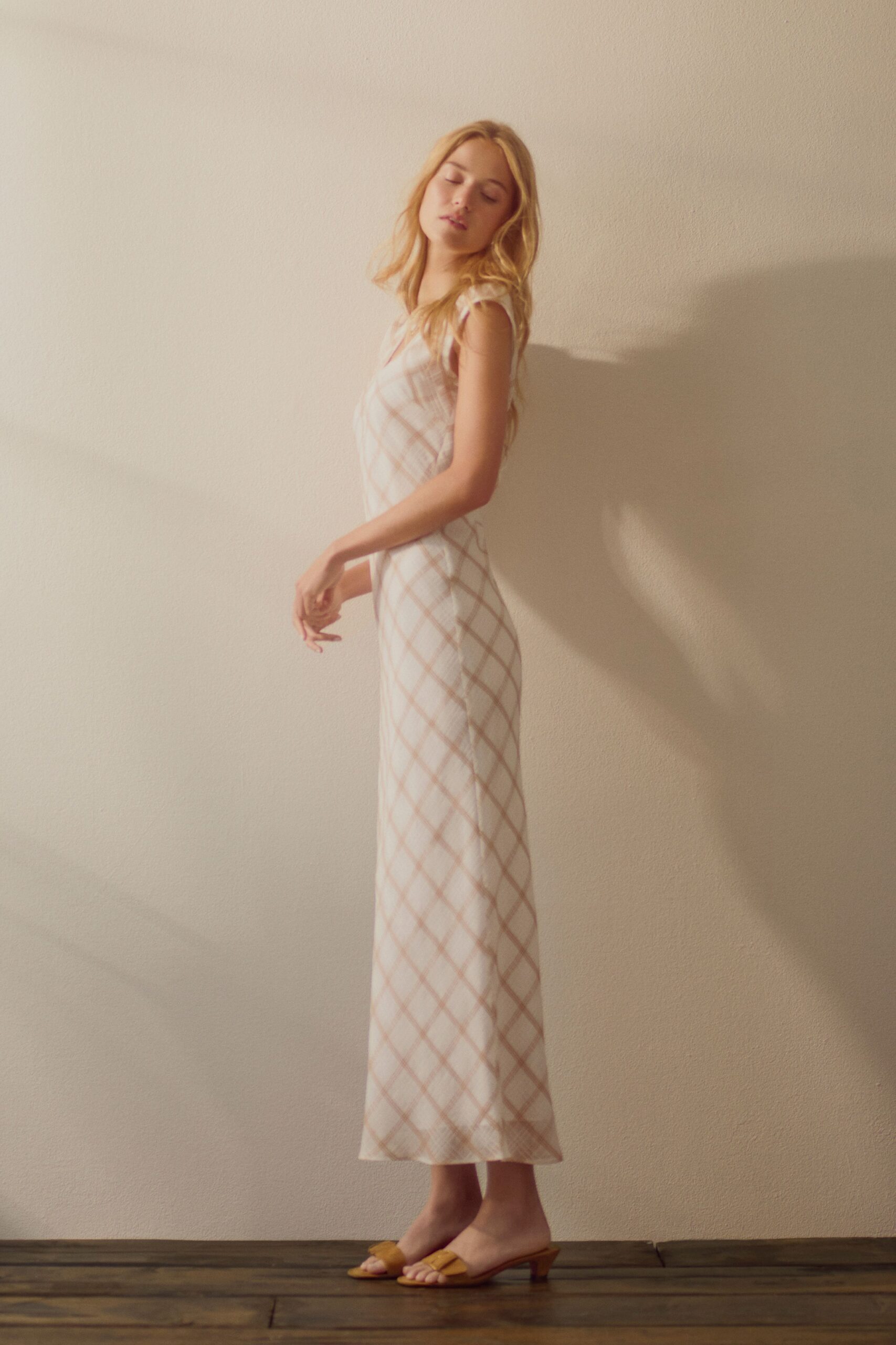 Bias-cut ivory slip dress in blush windowpane plaid, cap sleeves barely fluttering. Butterscotch leather mules with a folded vamp knot and low kitten heel. Sun warming the motel wall before checkout gets mentioned.