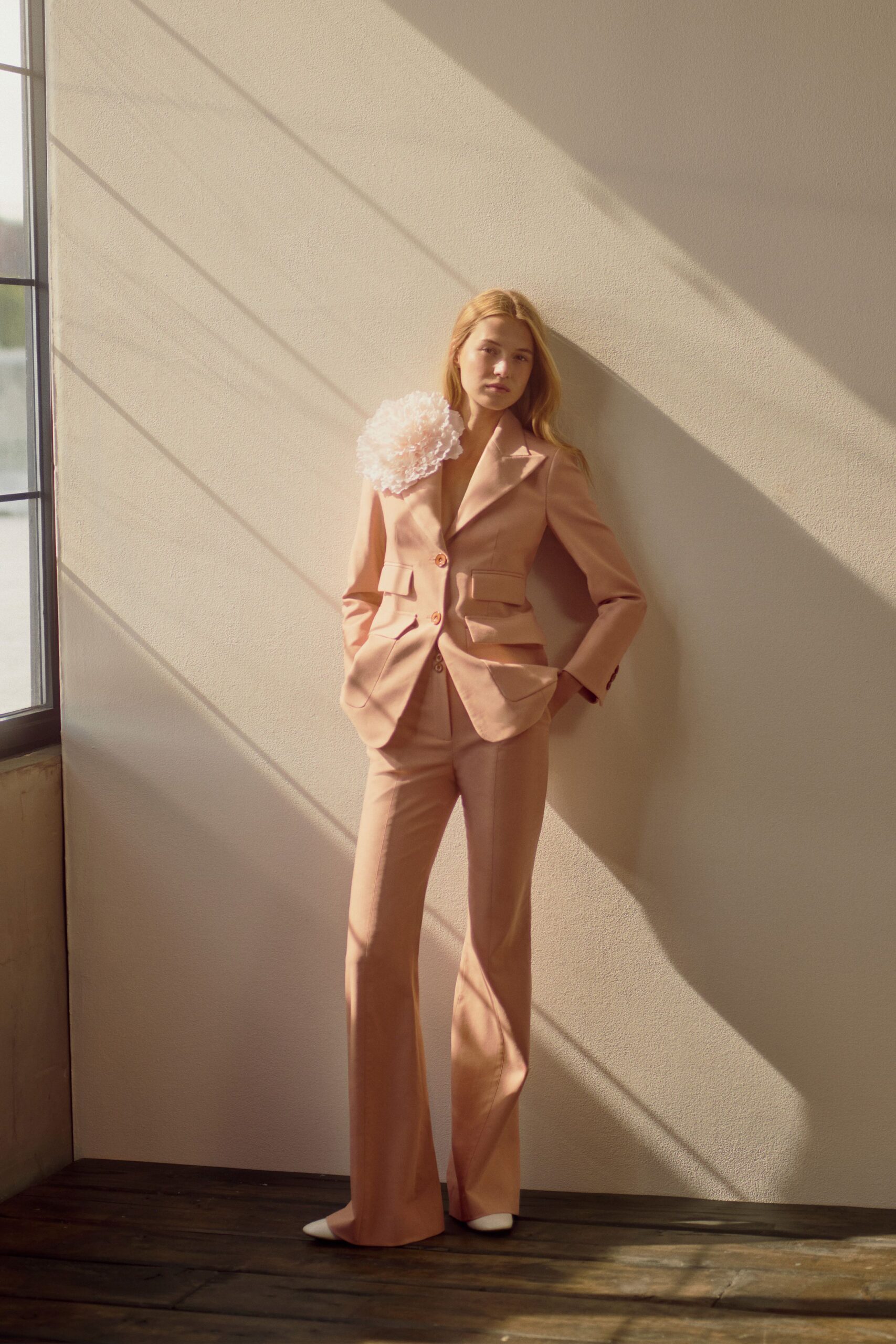 Powder-blush tailored blazer with jutting flap pockets and a single flowered shoulder. Matching flared trousers with a pressed crease and long break. White pointed shoes disappearing under the hem. Afternoon light in an empty townhouse before the ceremony starts.