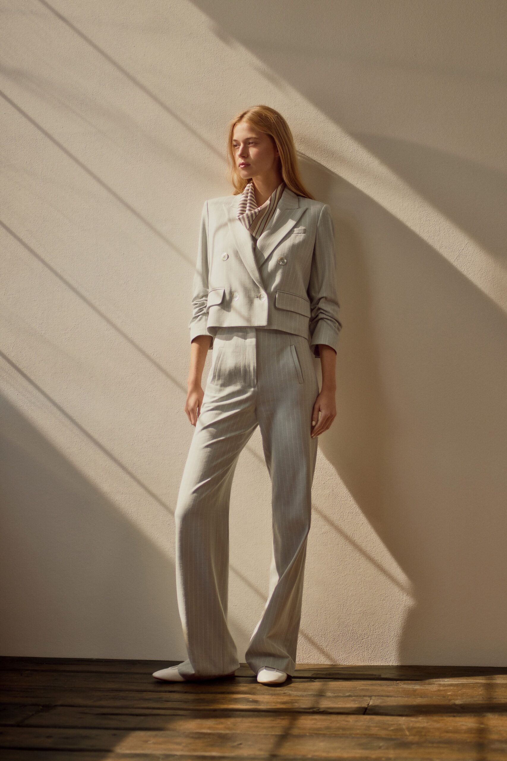 Cream and mauve striped scarf tucked into the neckline. Cropped dove-grey pinstripe jacket with pearly double-breasted buttons. High-waisted matching trousers, long break pooling softly over the shoes. White pointed flats. Sunlit hotel corridor before the conference rooms open.