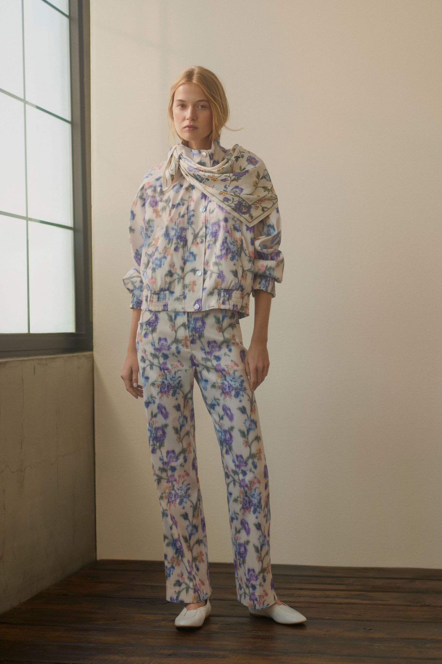 Cream silk scarf printed with violet wildflowers, folded square under the chin. Boxy floral jacket with pearly snaps and shirred hem and cuffs. Straight ivory trousers in the same blurred garden print. White square-toe flats. Sun through old studio glass, picking up flowers from the corner market.