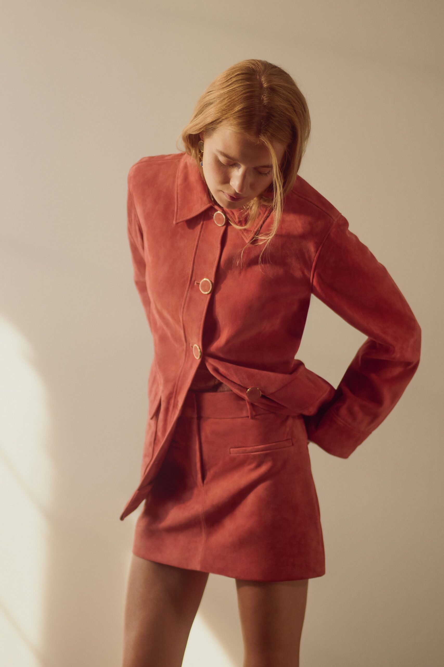 Terracotta suede jacket with a pointed collar and pale rimmed buttons. Matching suede mini, welt pocket set low at the hip. Small drop earrings with a blue glint. Late afternoon apartment heat, deciding whether to leave the windows open.